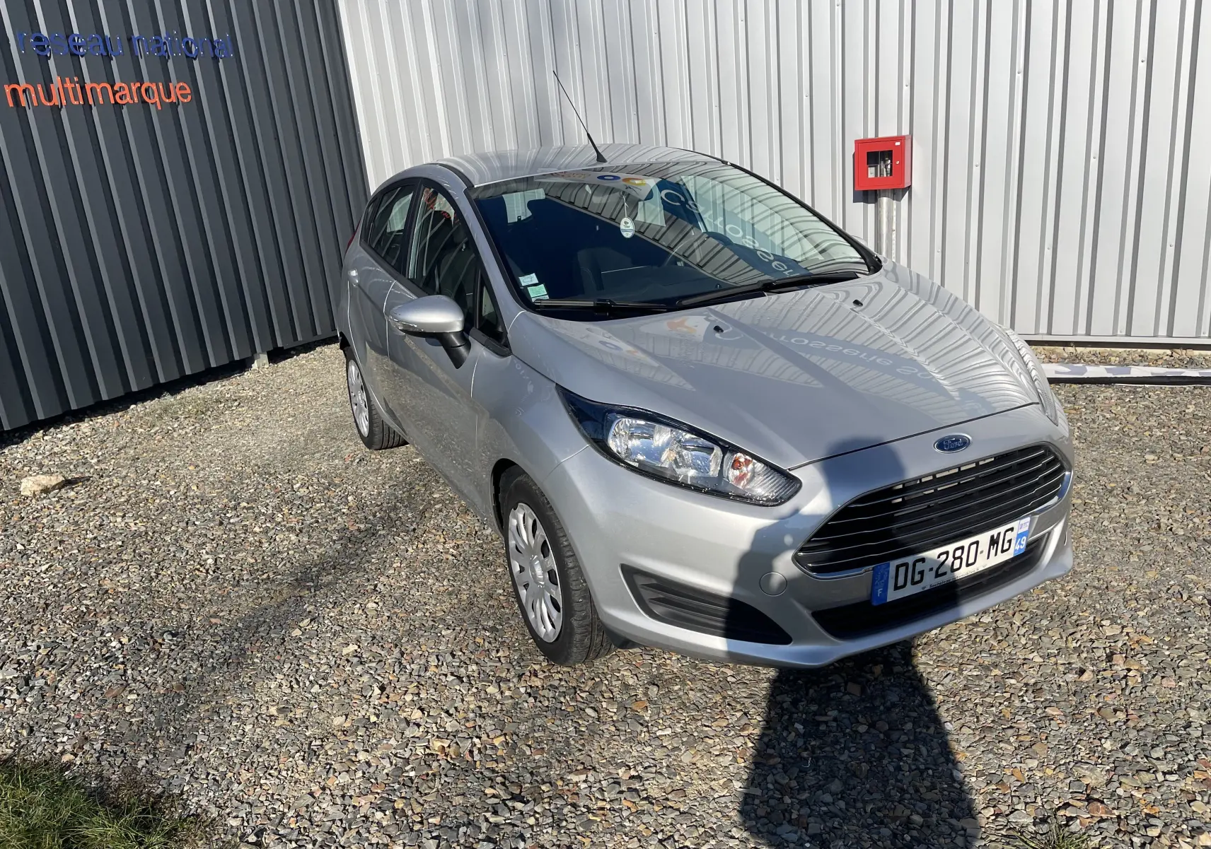 Ford Fiesta gris titane vue en 3/4 avant droit, garée sur sol gravillonné devant un mur métallique.