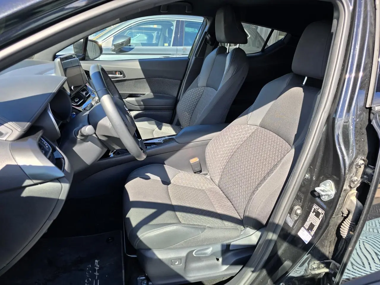 Vue intérieure côté conducteur du Toyota C-HR 2023 noir métal, sièges tissu/cuir avec surpiqûres grises et console centrale moderne.