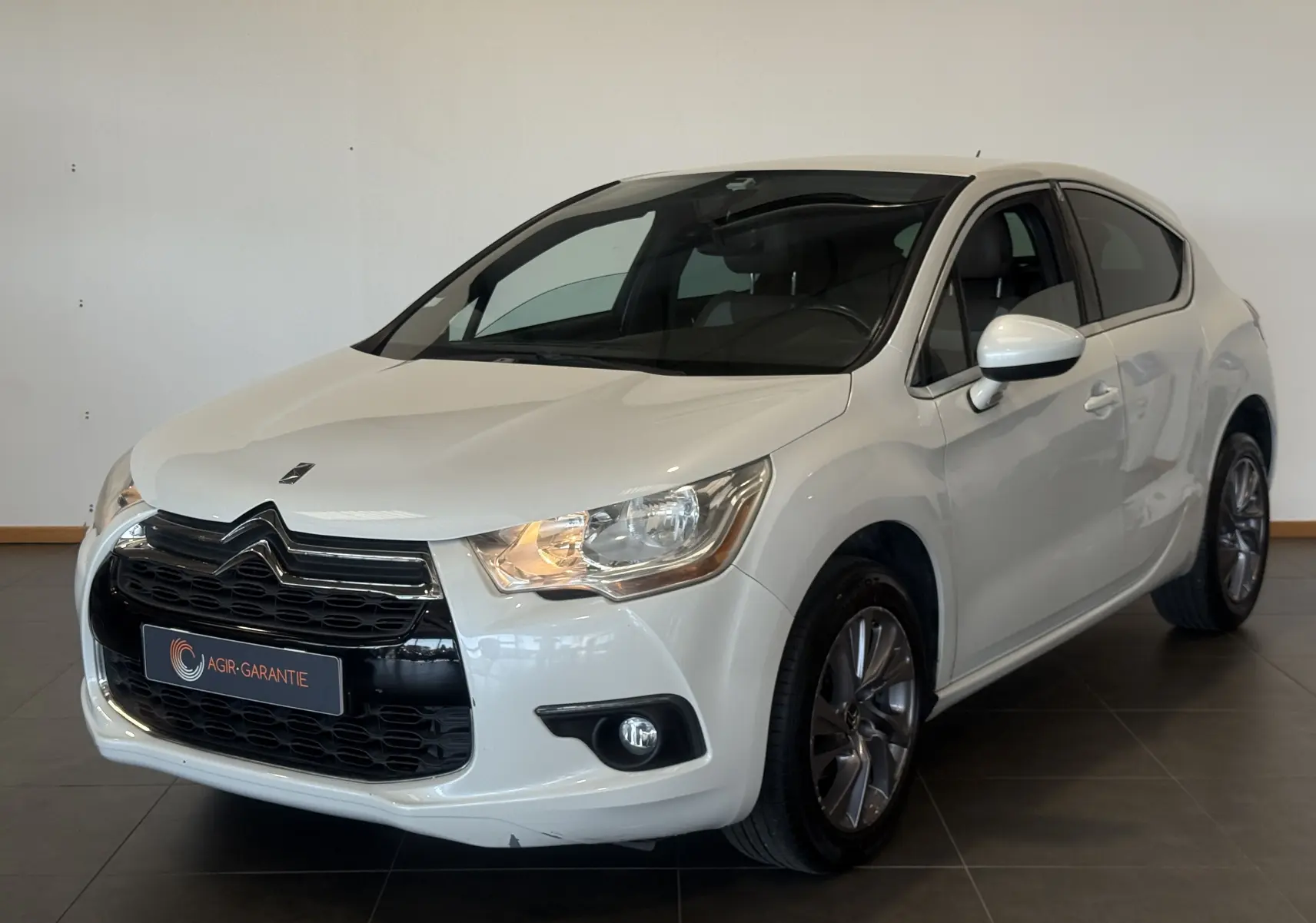 Citroën DS4 blanc vue 3/4 avant droit, avec calandre noire distinctive et phares allumés dans un intérieur neutre.