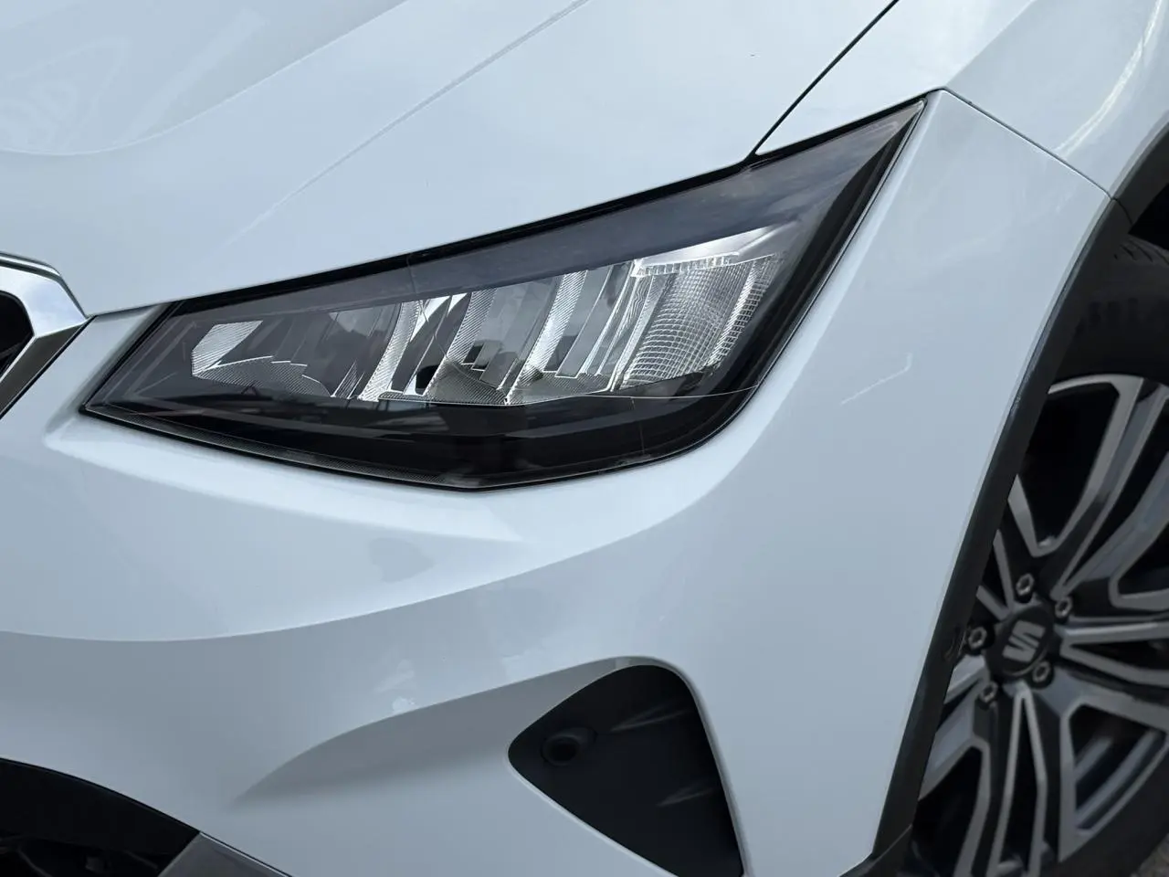 Gros plan sur le phare avant droit et la roue alliage 17'' d'un SEAT Arona blanc Nevada finition Xpérience 2025.