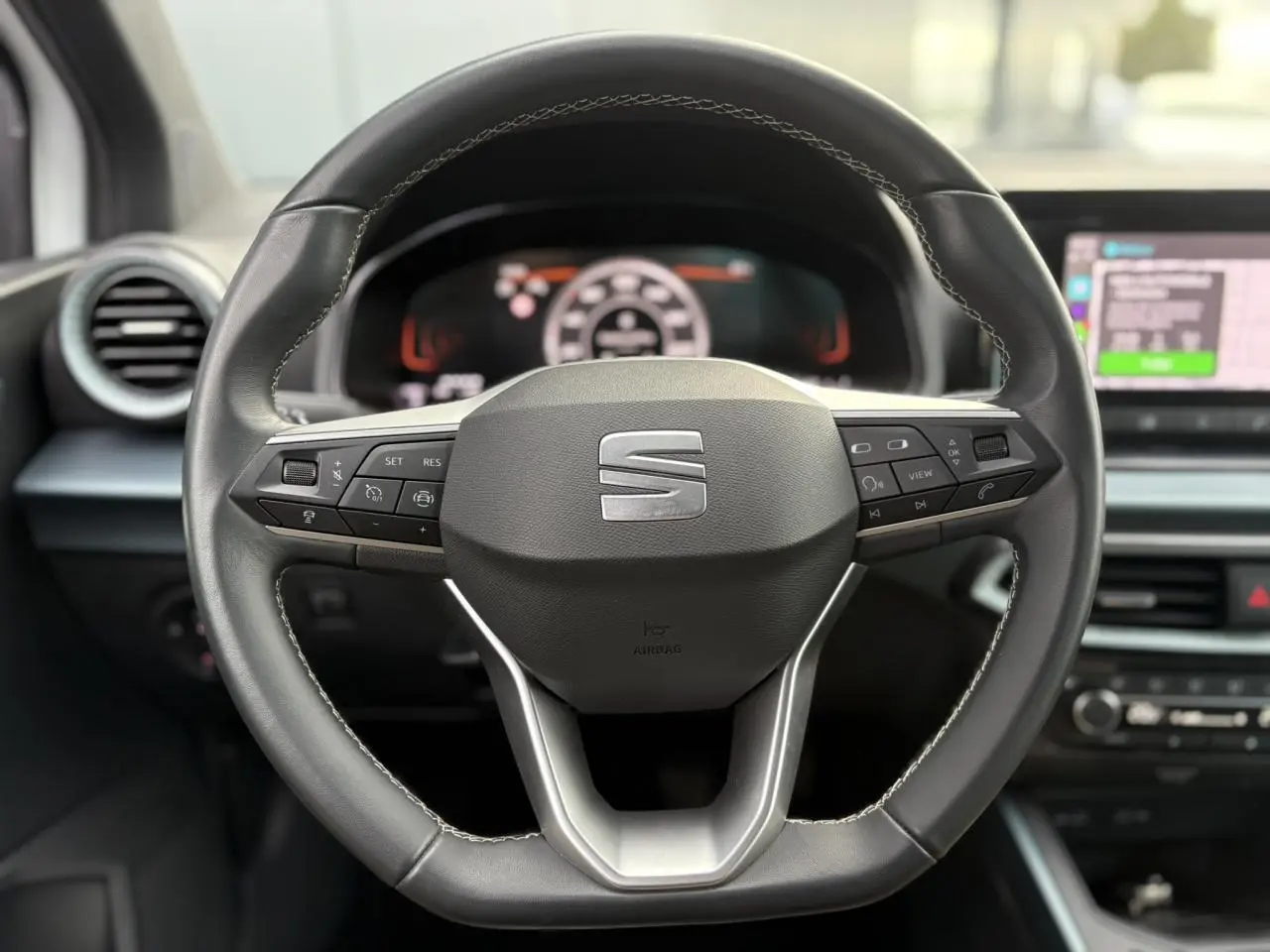 Gros plan sur le volant cuir multifonction de la SEAT Arona 2025 avec commandes intégrées et tableau de bord numérique flou.