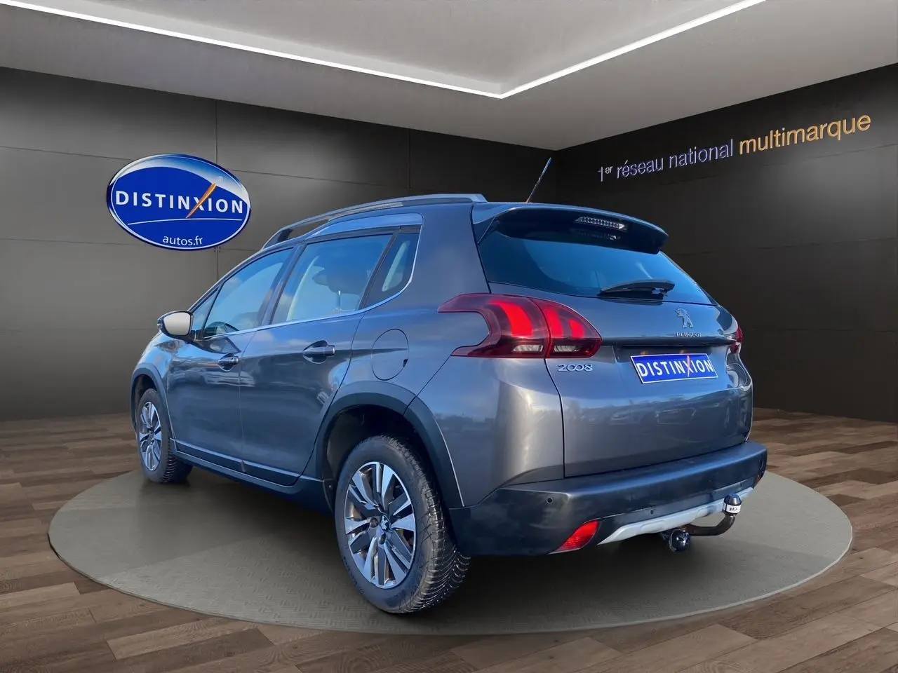 Vue 3/4 arrière droite d'un Peugeot 2008 gris platinium avec barres de toit et attelage visible en intérieur showroom.