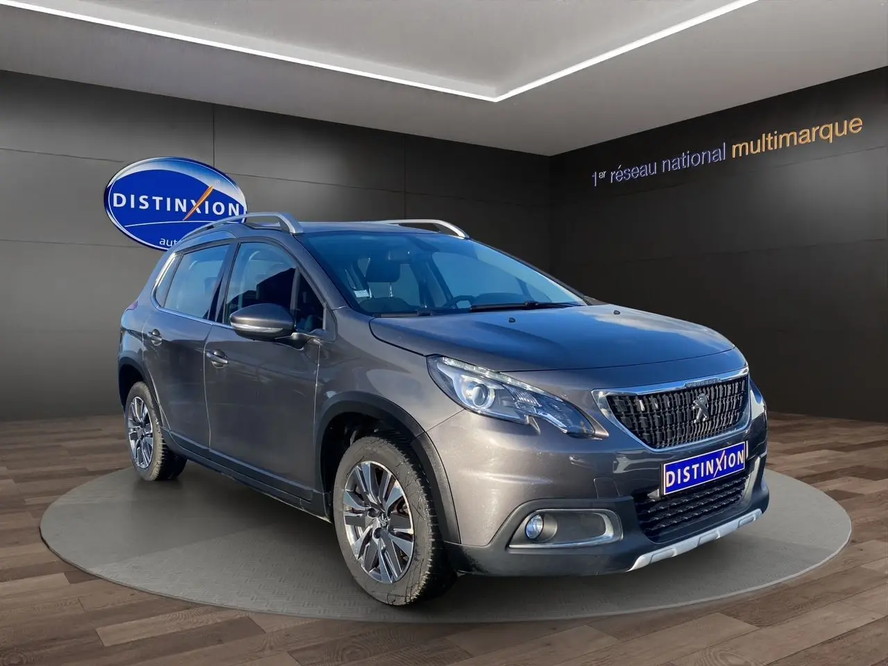 Peugeot 2008 gris platinium en 3/4 avant droit, avec jantes alliage et barres de toit visibles en showroom.