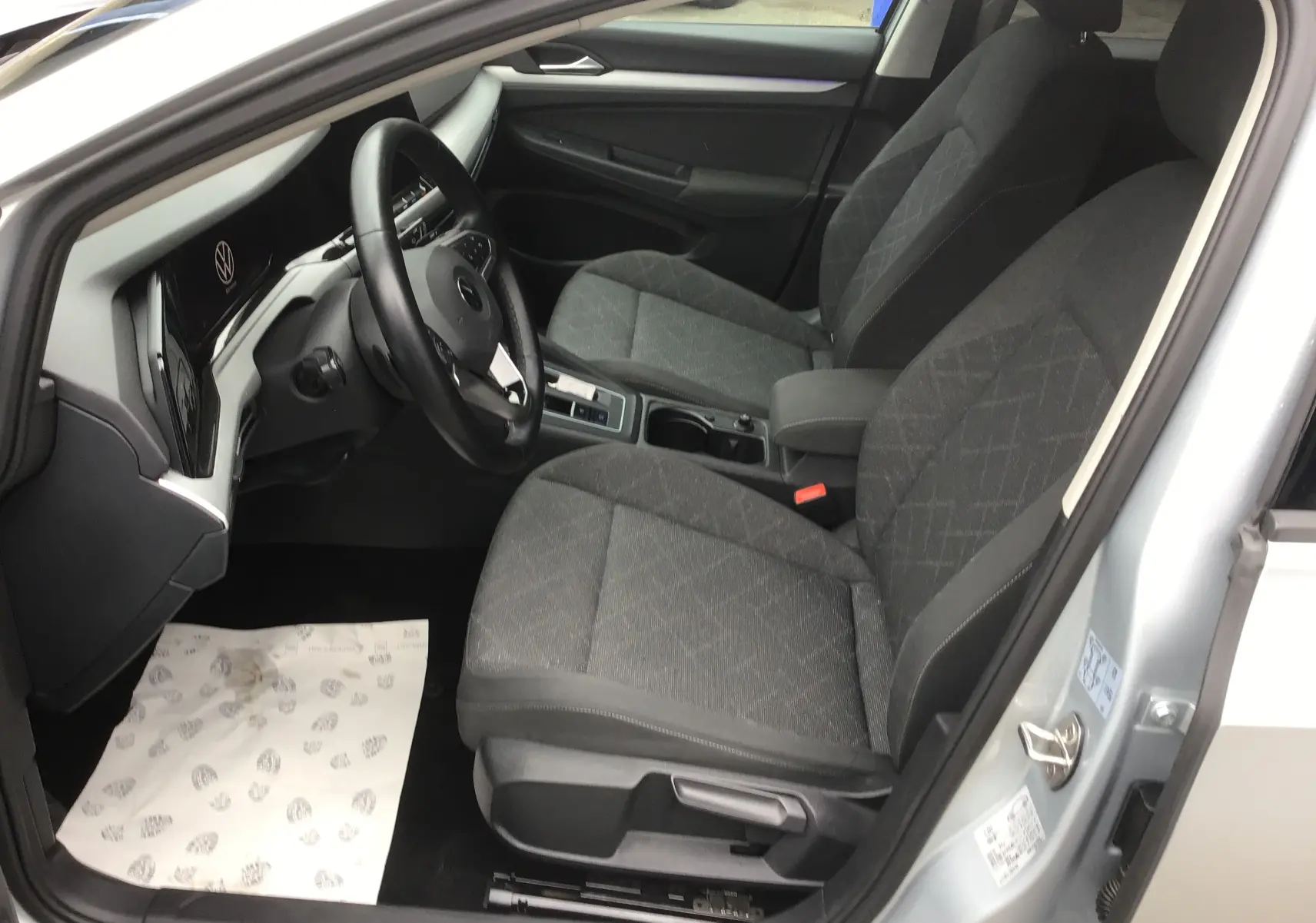 Vue intérieure côté conducteur de la Volkswagen Golf VIII gris reflex, montrant les sièges avant et le tableau de bord moderne.