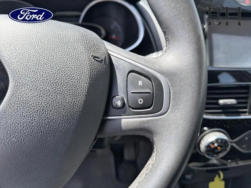 Gros plan sur le volant cuir noir avec commandes intégrées d’une Renault Clio blanche, tableau de bord partiellement visible.