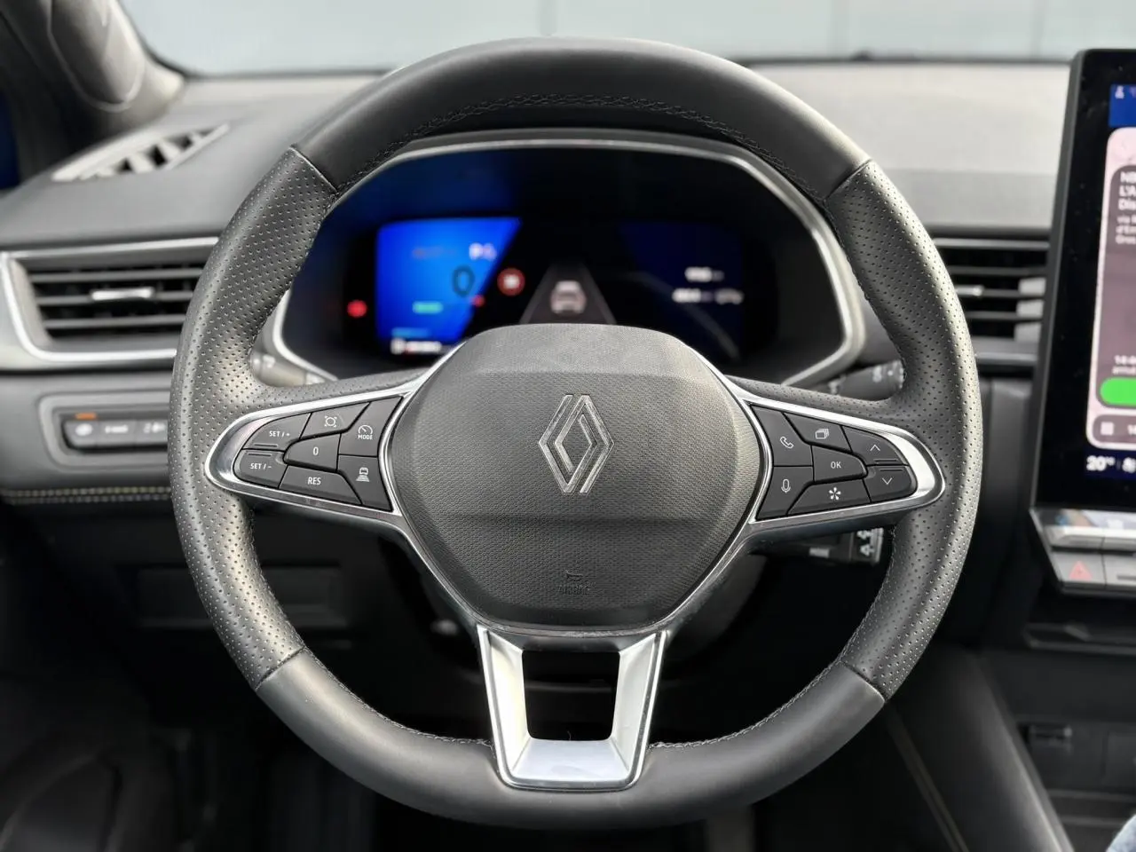 Vue rapprochée du volant noir perforé avec logo Renault, tableau de bord numérique et écran tactile à droite.