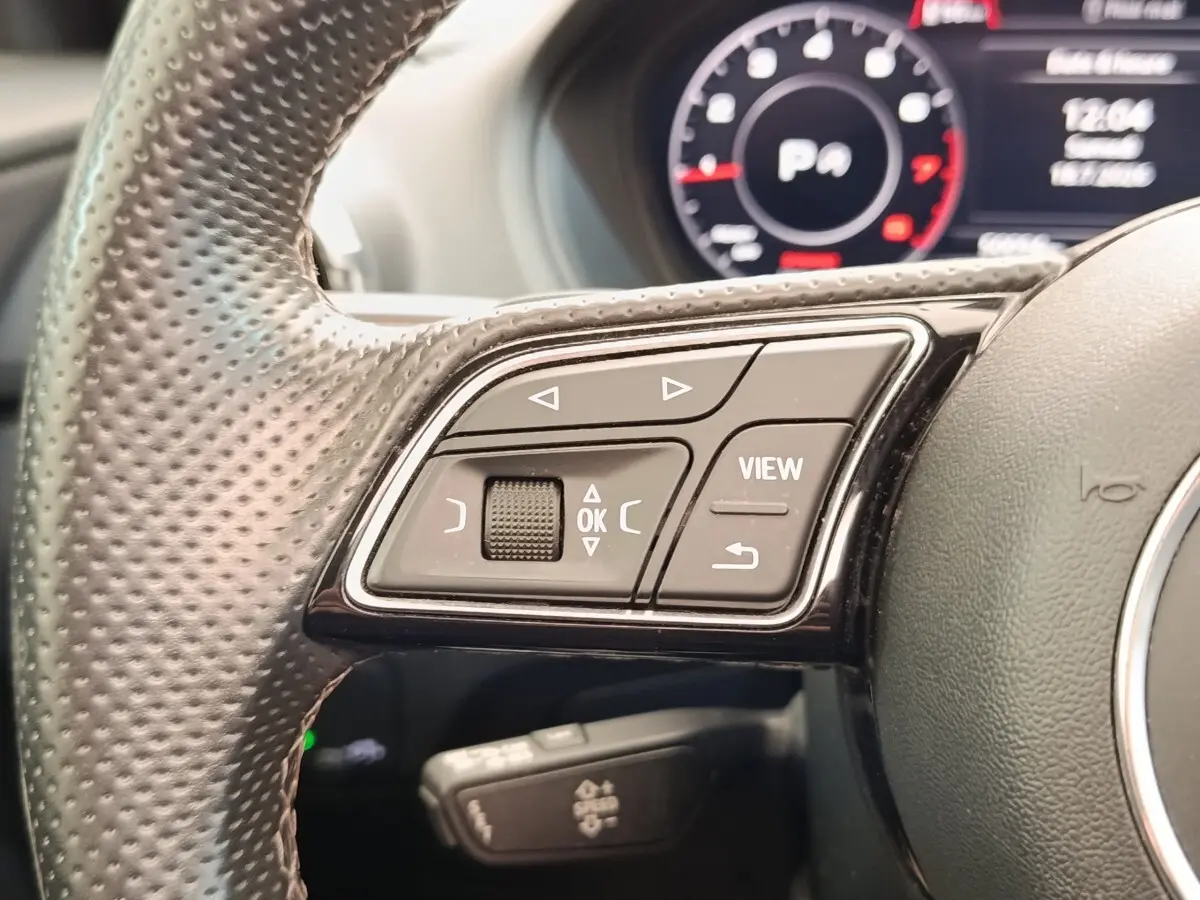 Gros plan sur les commandes gauche du volant en cuir perforé d’un Audi Q2 noir, avec tableau de bord numérique en arrière-plan.