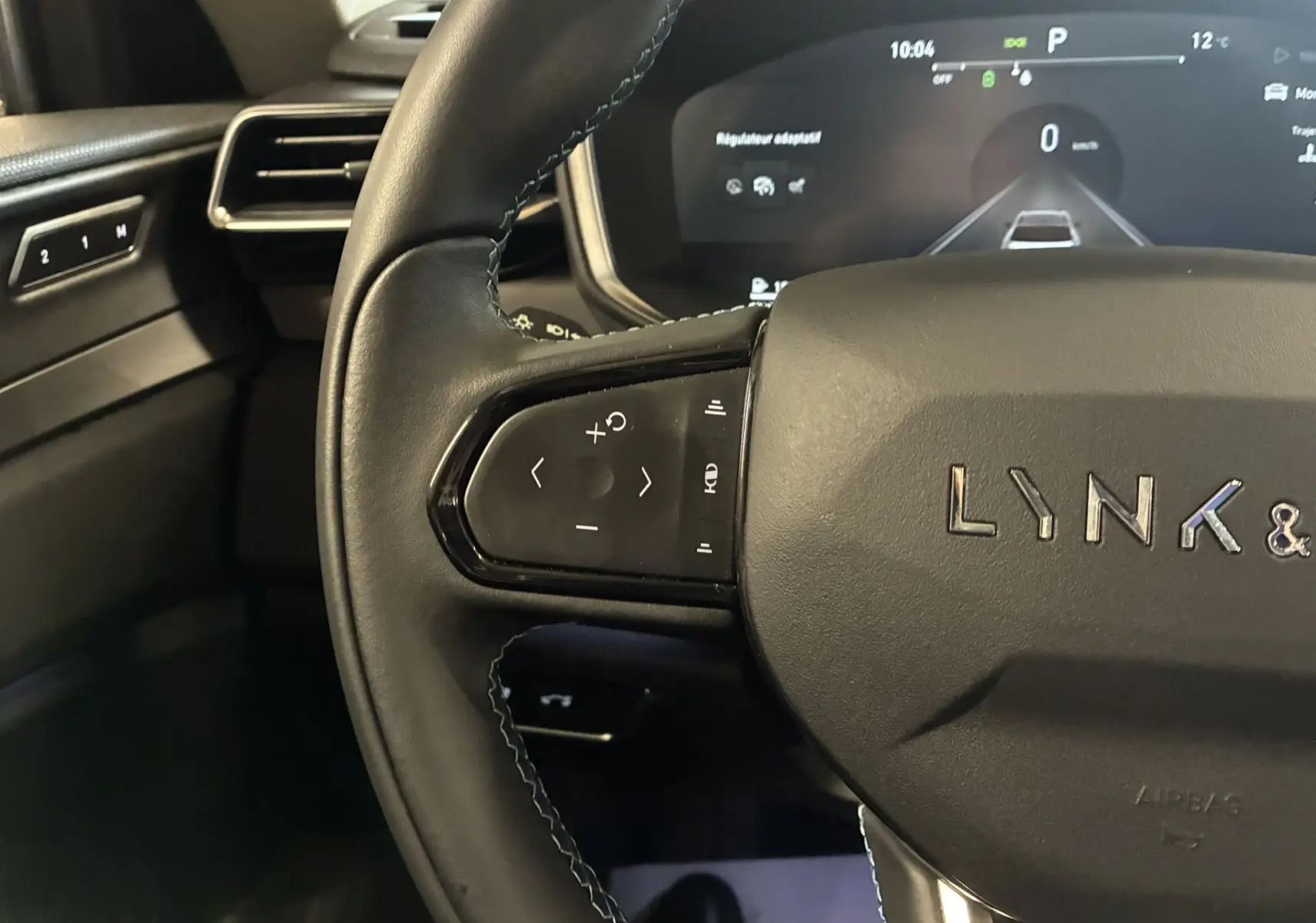 Vue rapprochée du volant noir de la LYNK & CO 01 avec commandes intégrées et tableau de bord digital en arrière-plan.