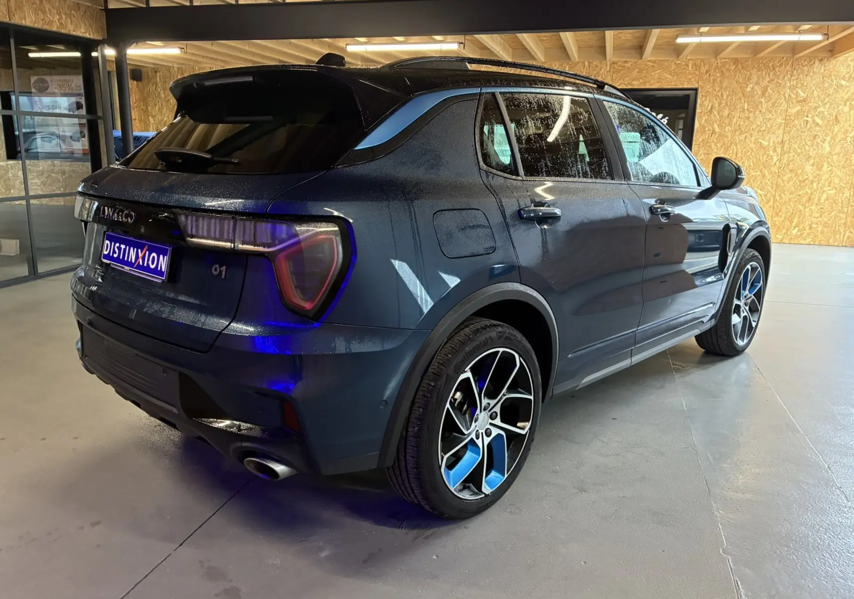 Vue 3/4 arrière droite du SUV Lynk & Co 01 bleu hybride rechargeable avec toit noir et jantes bicolores en showroom.