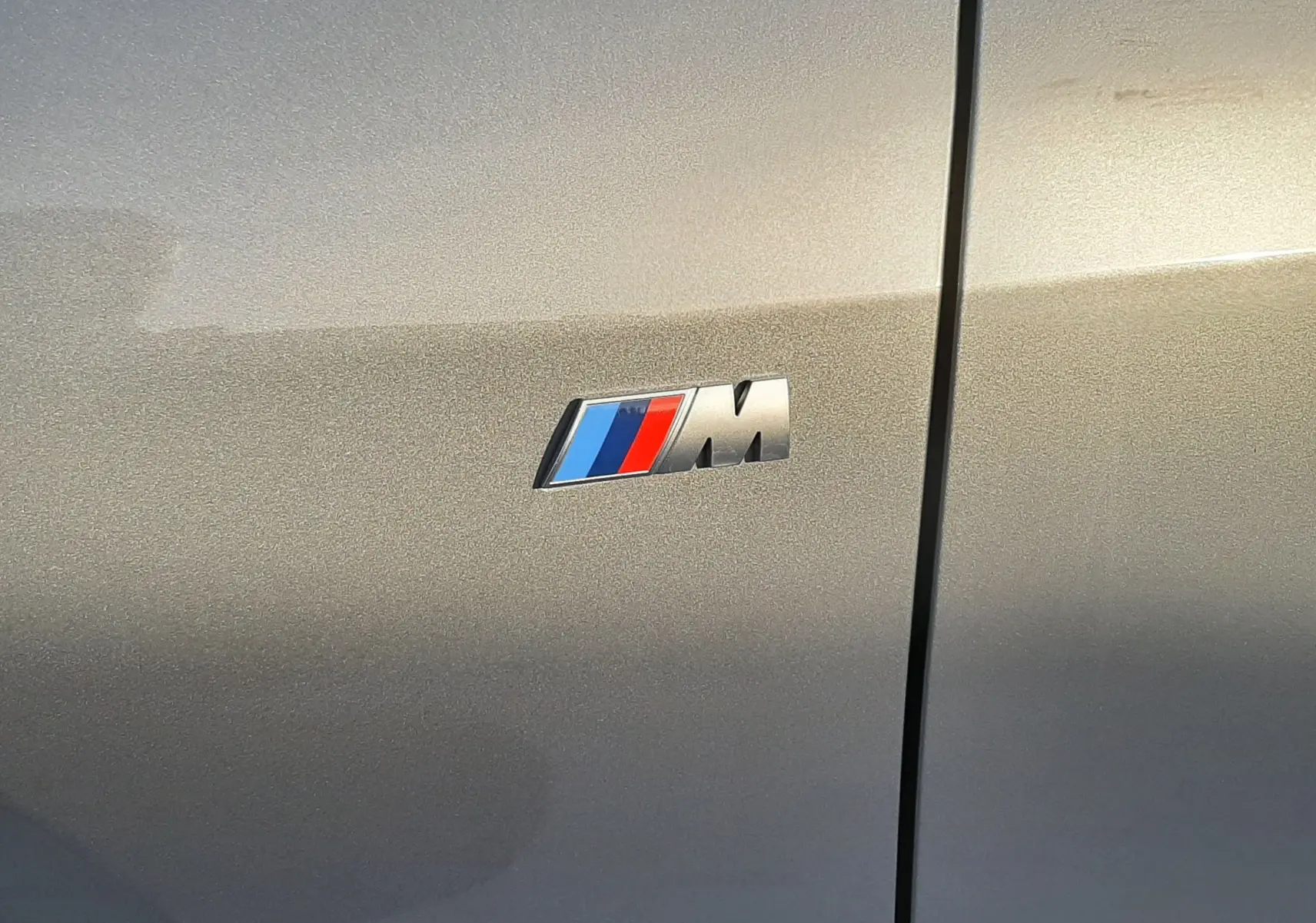 Gros plan sur l'emblème M sur la carrosserie gris Skyscraper d'une BMW Série 1 F70 120D M Sport.