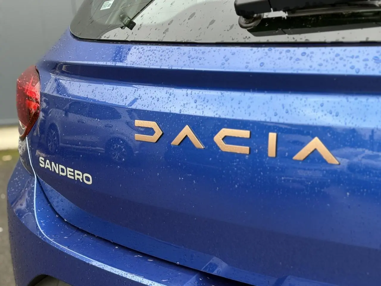 Gros plan arrière gauche sur le coffre bleu Iron de la Dacia Sandero avec lettrage cuivré sous la pluie.