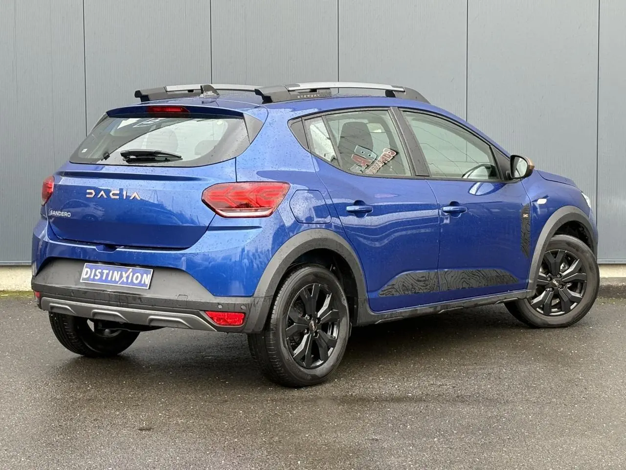 Vue 3/4 arrière droite d'une Dacia Sandero Stepway Bleu Iron avec jantes noires et barres de toit grises.