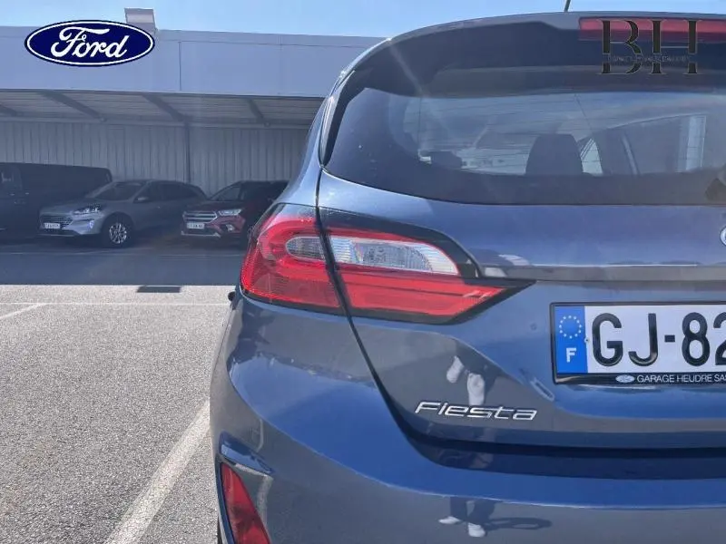 Vue arrière droite d'une Ford Fiesta bleu azur métallisée avec feux arrière et logo visibles sur un parking.
