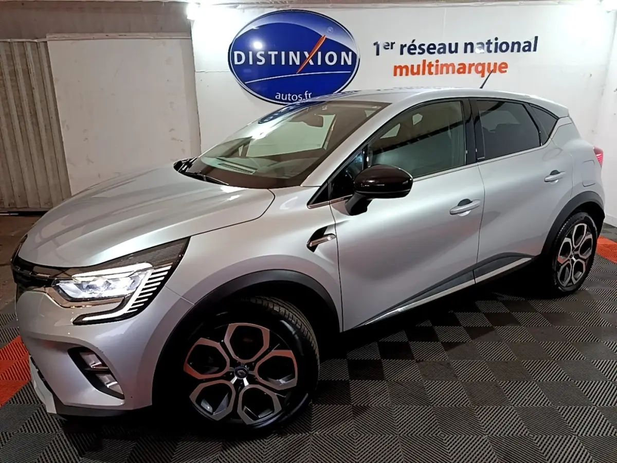 Renault Captur Intens E-Tech hybride rechargeable gris clair, vue 3/4 avant droit avec jantes distinctives et feux LED allumés.