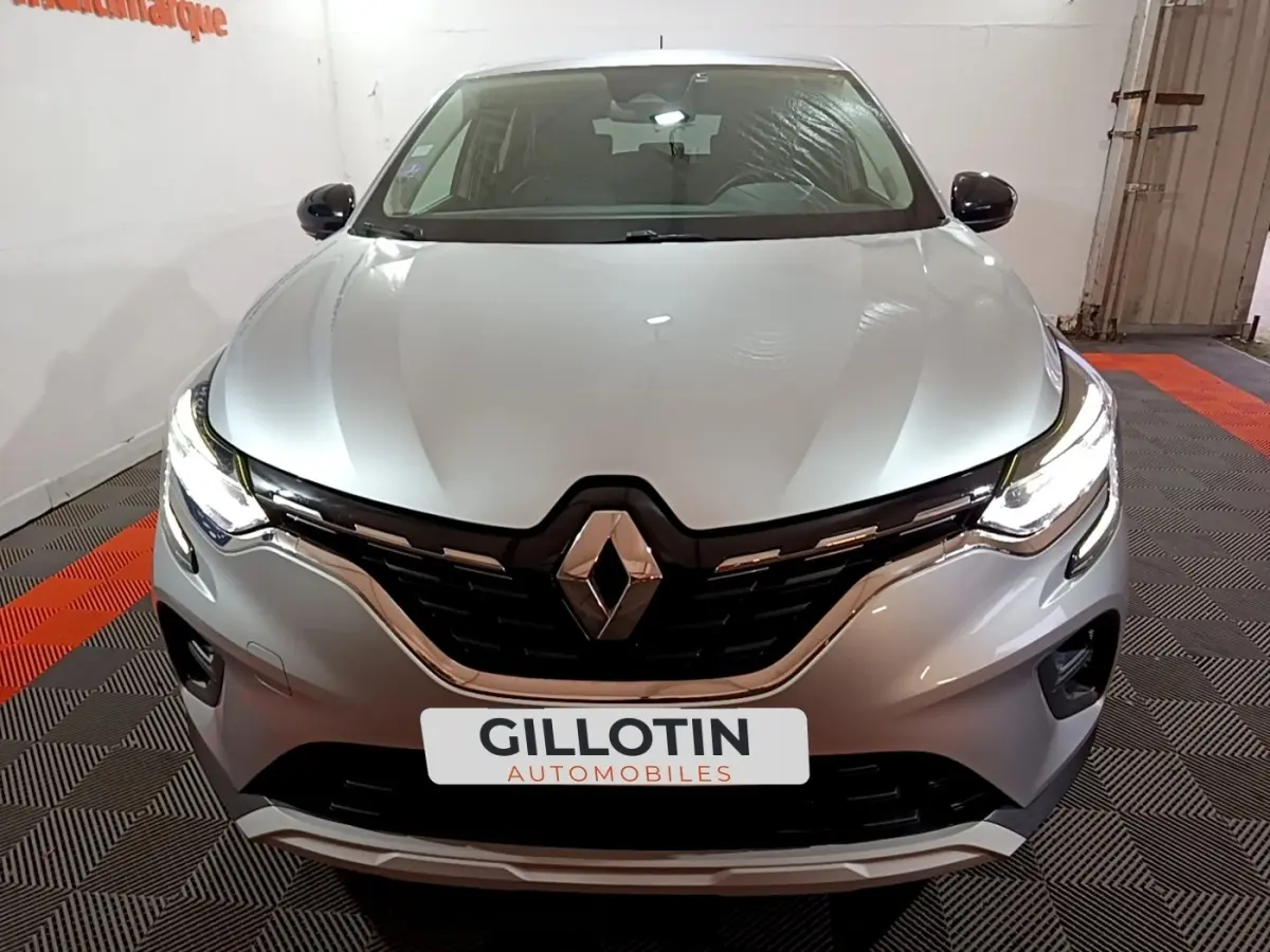 Vue avant d'un Renault Captur Intens E-Tech hybride rechargeable gris clair avec phares LED allumés en intérieur showroom.