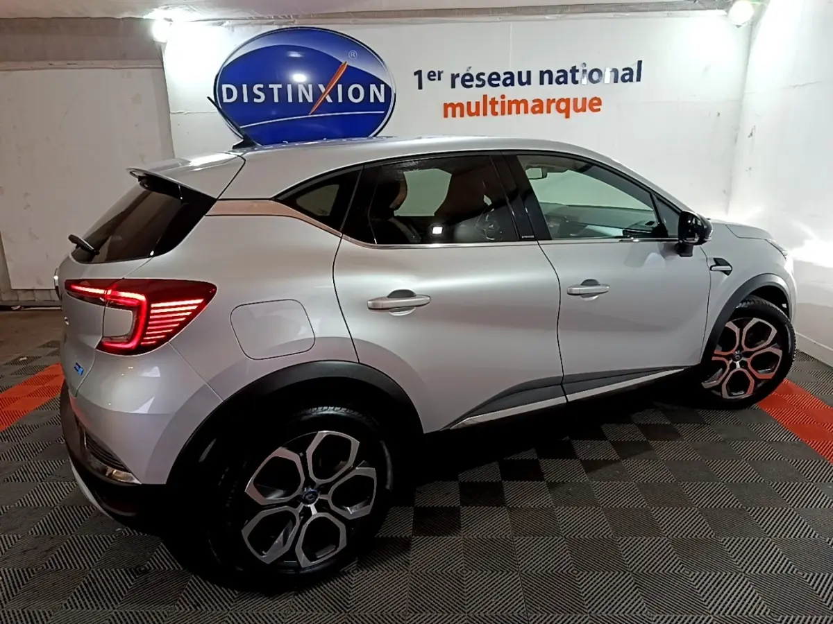 Vue latérale droite d’un Renault Captur Intens E-Tech hybride rechargeable gris clair avec feux arrière LED allumés en intérieur showroom.