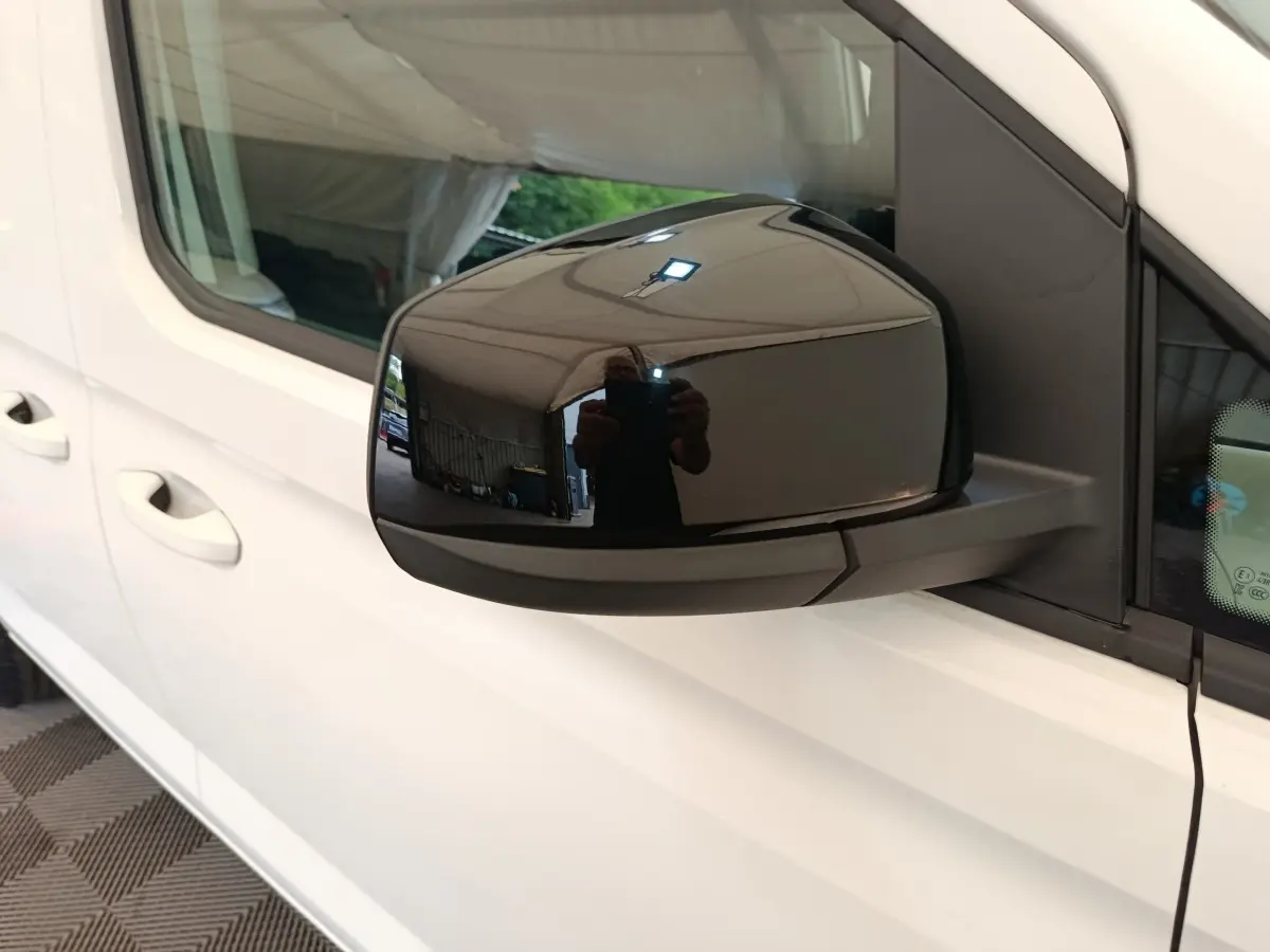 Rétroviseur noir côté conducteur d’un Volkswagen Caddy blanc vu de près, avec reflet de l’environnement intérieur.
