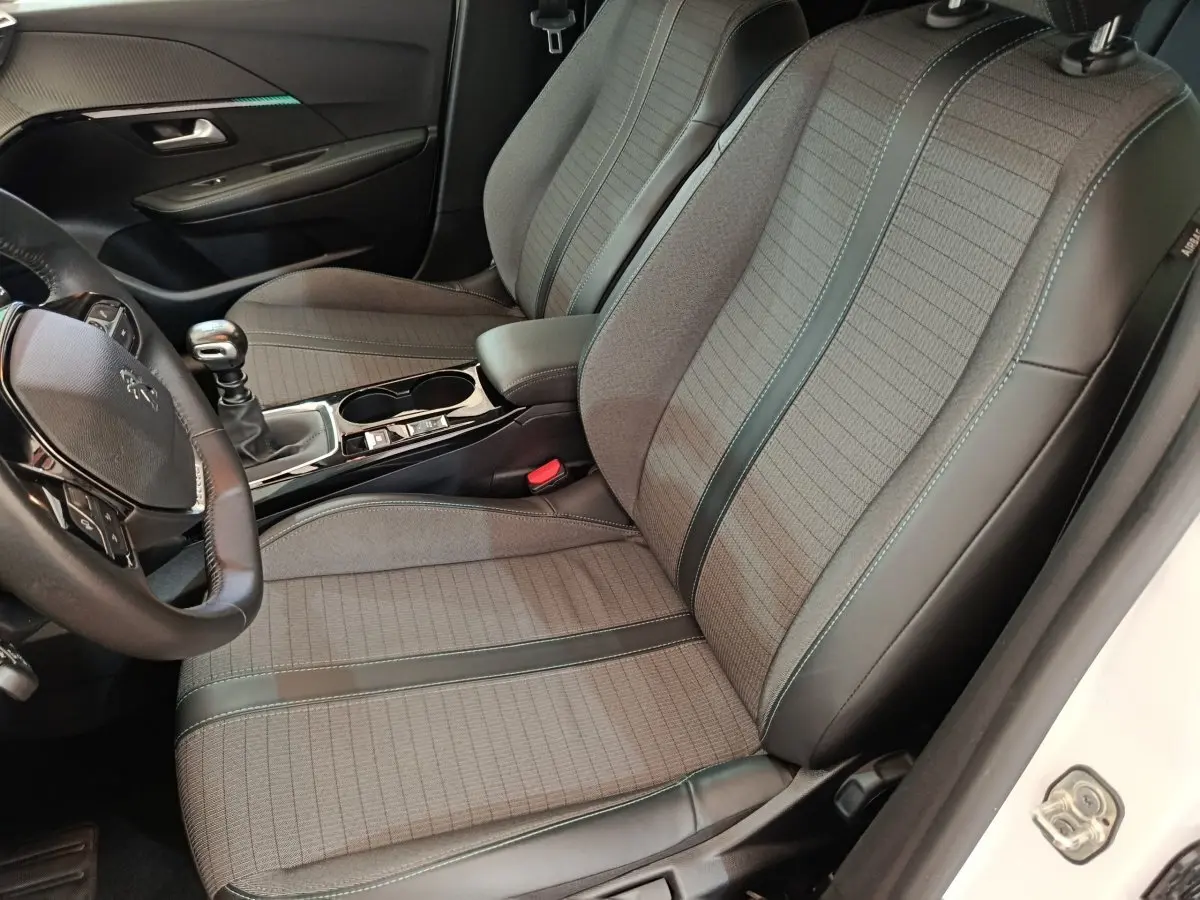 Vue intérieure côté conducteur de la Peugeot 208 blanche, montrant les sièges gris tissu avec surpiqûres et volant cuir multifonctions.