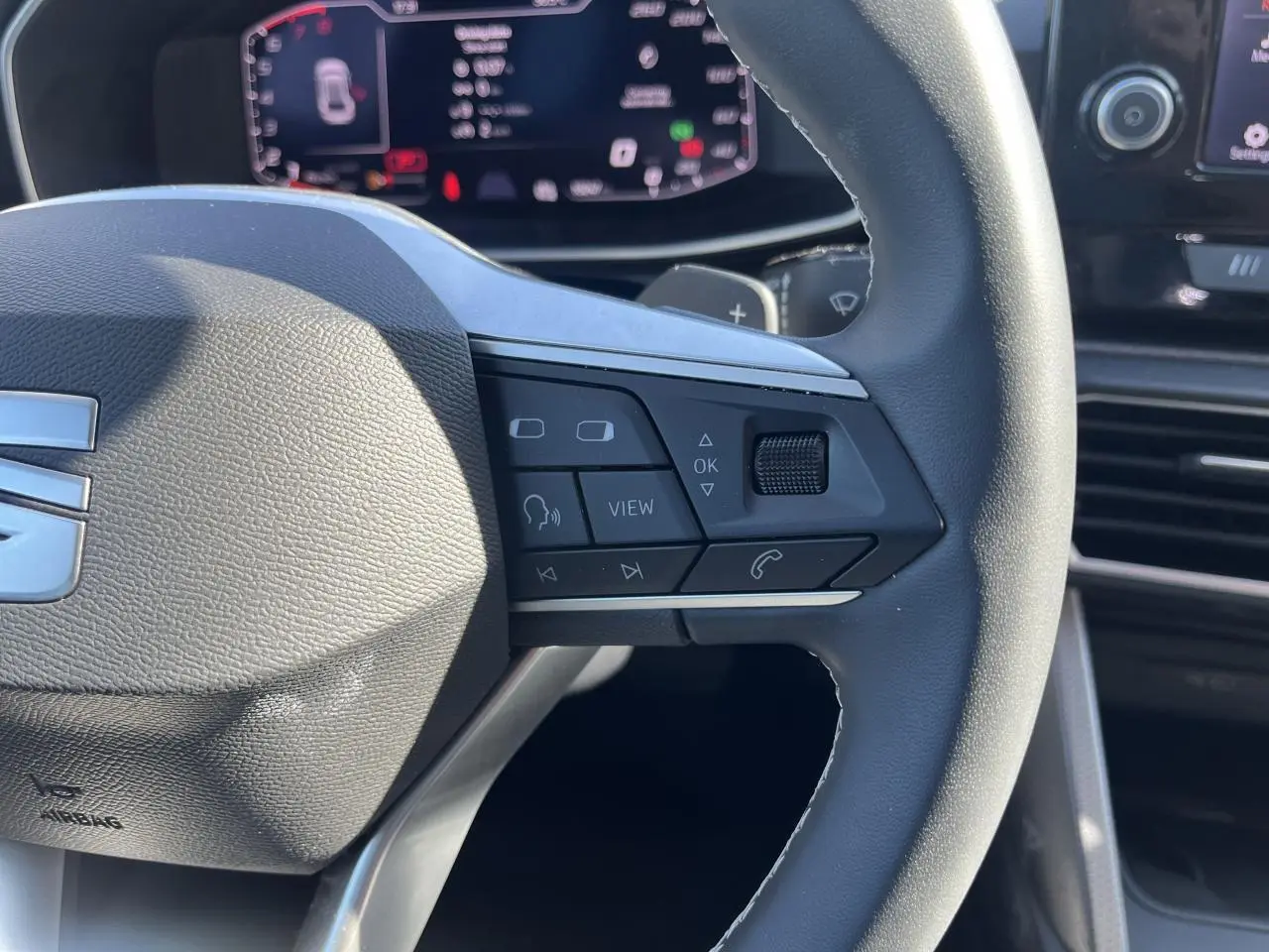 Gros plan sur le volant cuir multifonction de la SEAT LEON IV 2023 avec commandes vocales et écran digital en arrière-plan.