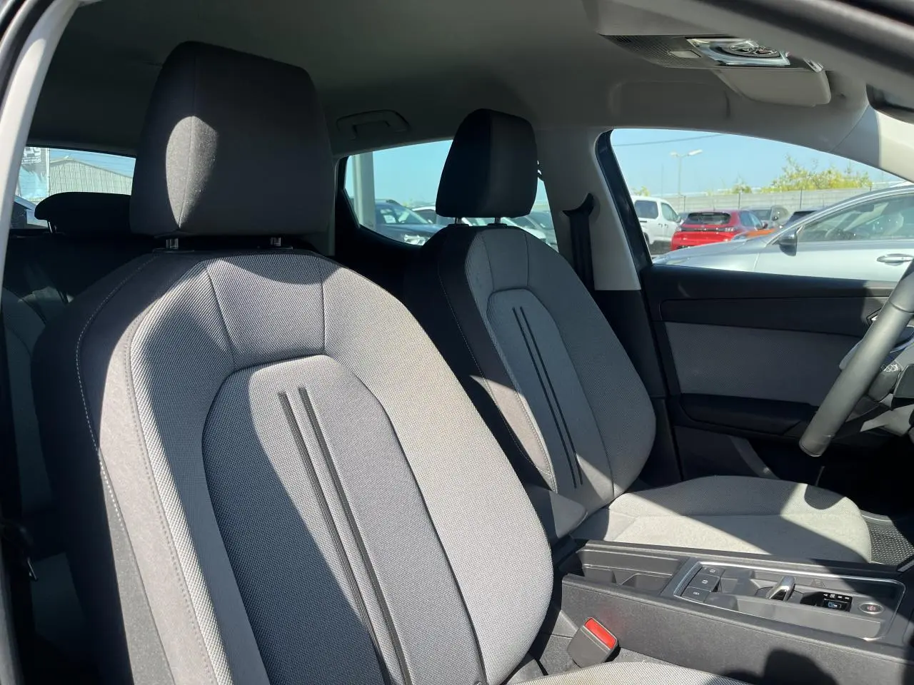 Intérieur avant de la SEAT LEON IV gris Magnetic Metal, sièges tissu gris et accoudoir central visible au soleil.