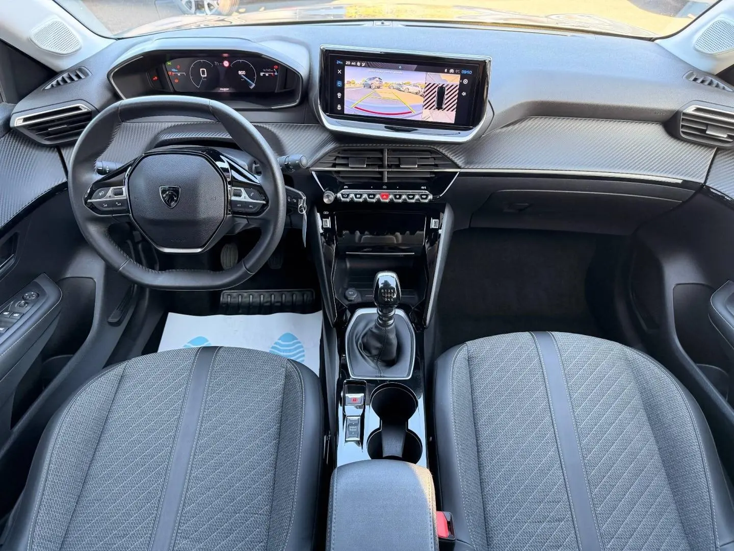 Vue intérieure avant de la Peugeot 208 1.2 essence 2025, volant compact, écran tactile et sièges tissu gris.