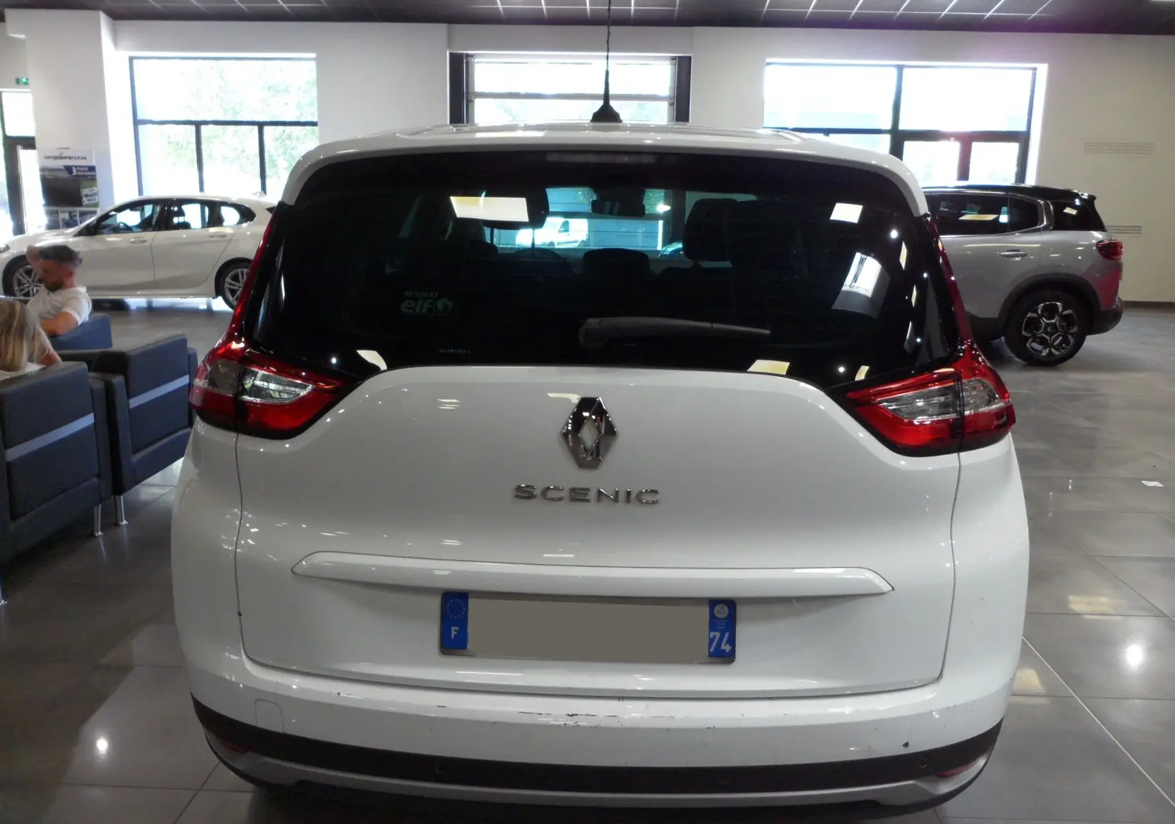 Vue arrière d'un Renault Grand Scenic IV blanc Glacier en intérieur, avec feux arrière rouges et logo Renault visible.