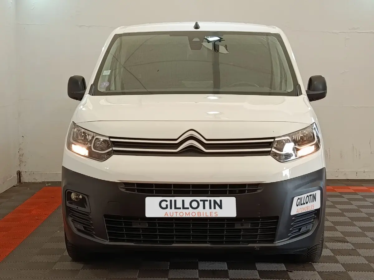 Vue avant d'un Citroën Berlingo blanc avec calandre noire et phares allumés dans un garage.