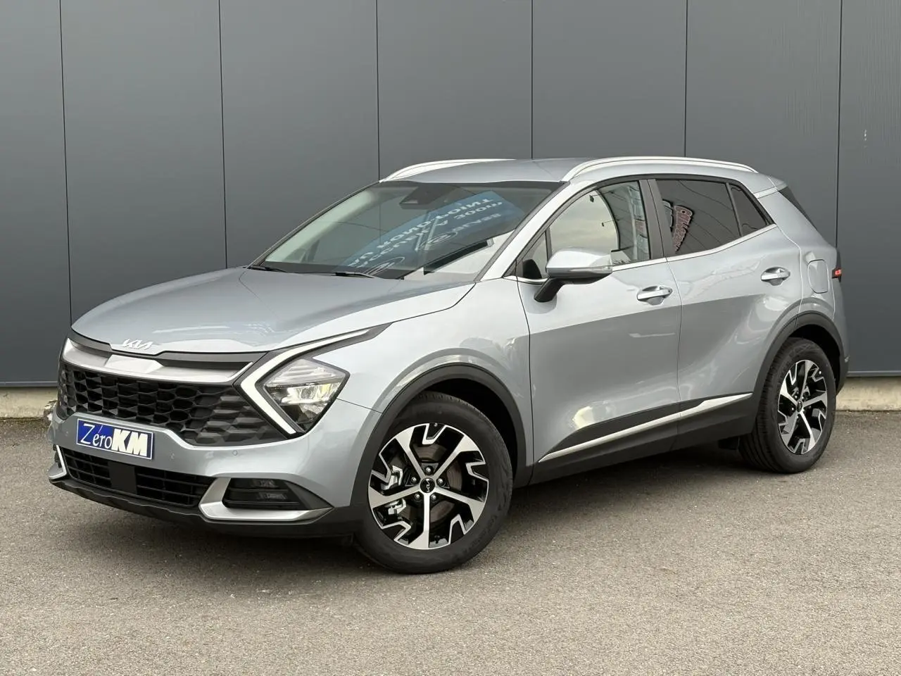 Kia Sportage gris perle en 3/4 avant droit, avec jantes alliage 18 pouces et calandre noire distinctive.