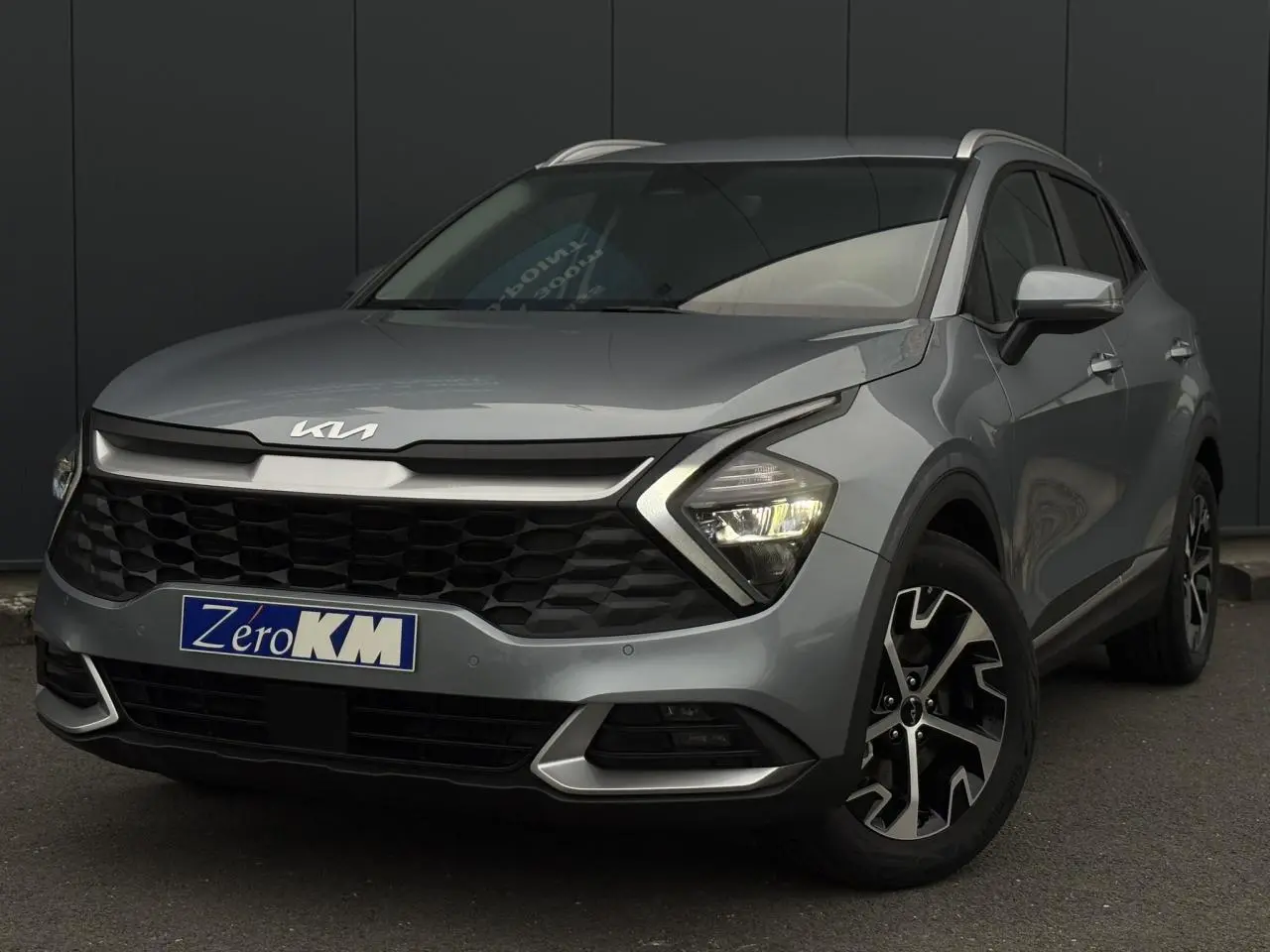 Kia Sportage gris perle en 3/4 avant droit, avec calandre noire et jantes alliage 18 pouces distinctives.