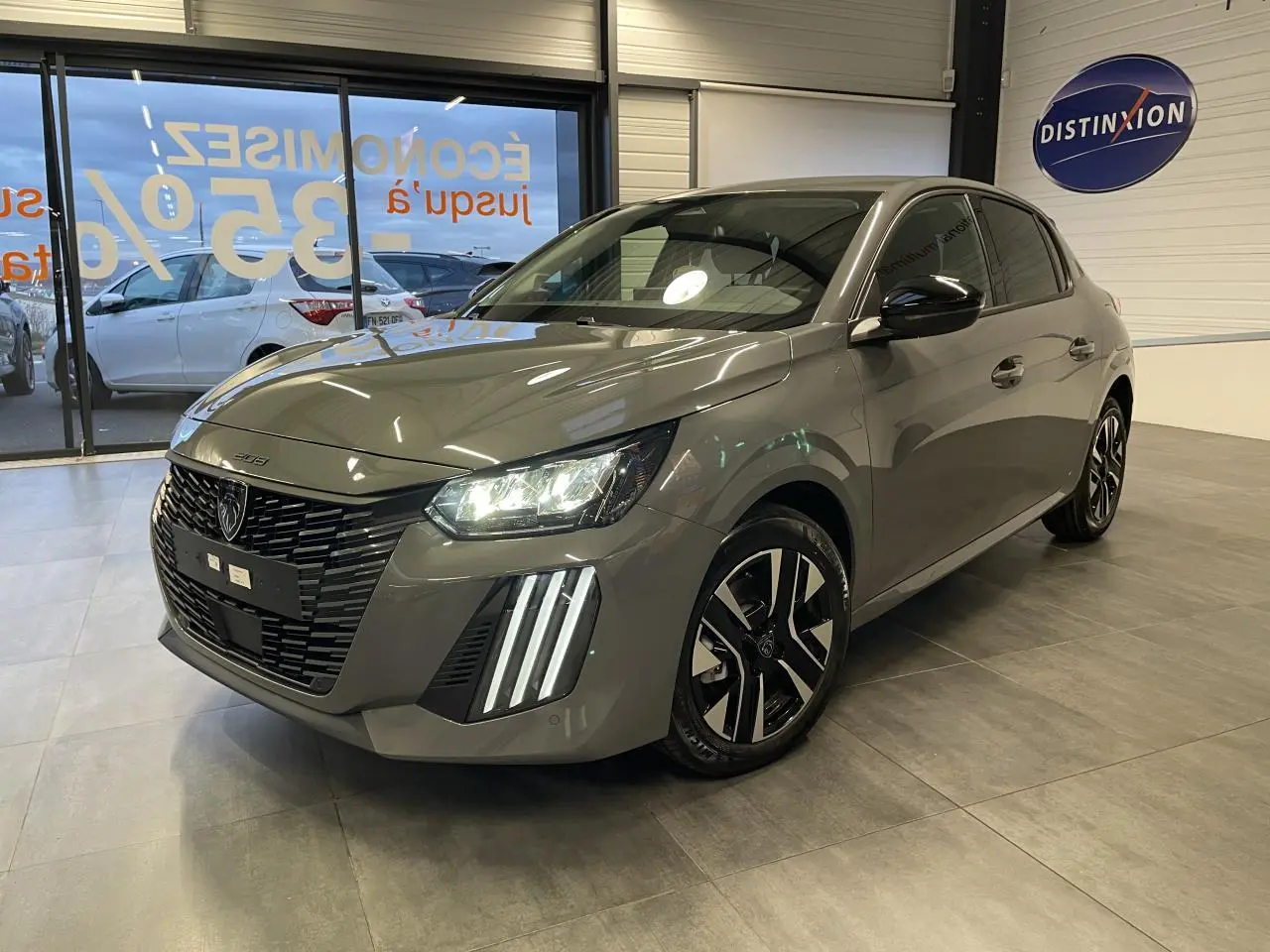 Peugeot 208 gris selenium vue 3/4 avant en intérieur, avec calandre noire et feux LED distinctifs allumés.