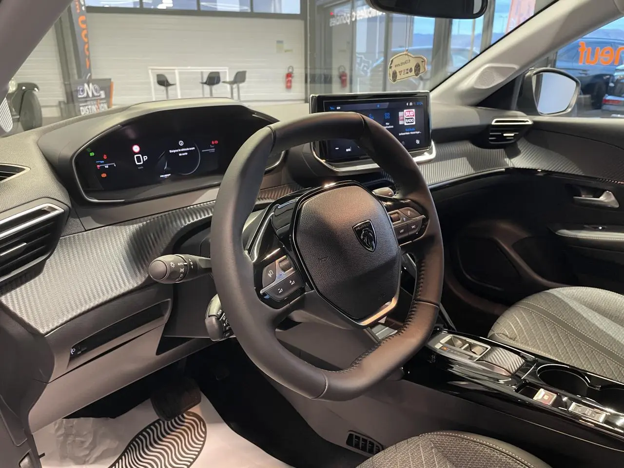 Vue intérieure de la Peugeot 208 2025 montrant le volant compact, tableau de bord numérique et console centrale noire brillante.