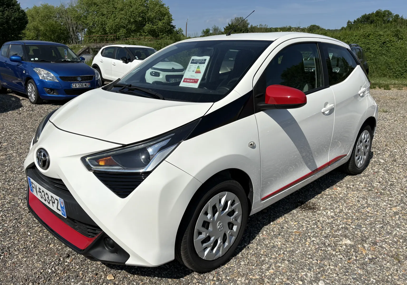 Toyota Aygo MC blanc vue 3/4 avant droit avec rétroviseurs rouges et bande rouge sur bas de caisse, en extérieur.