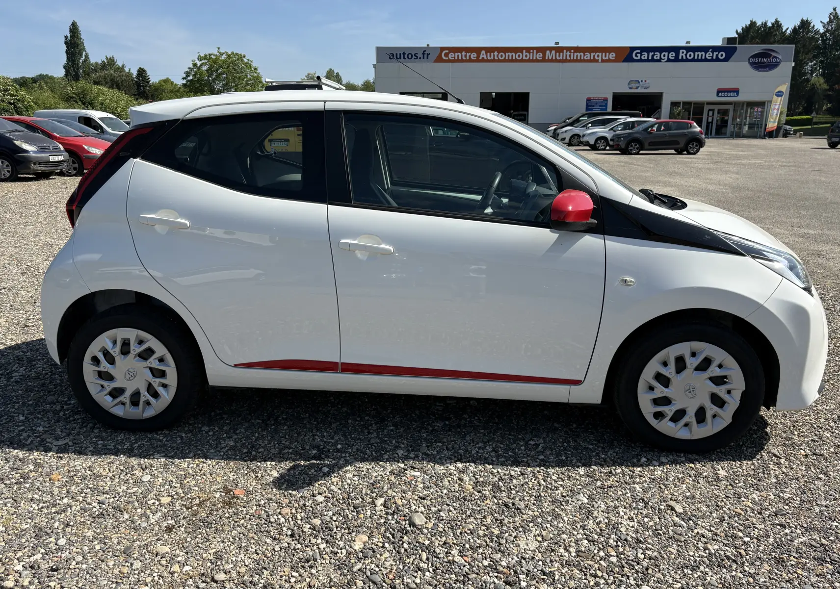 Profil droit d'une Toyota Aygo MC blanche 2020 avec rétroviseur et bandeau latéral rouges, stationnée sur un parking gravillonné.