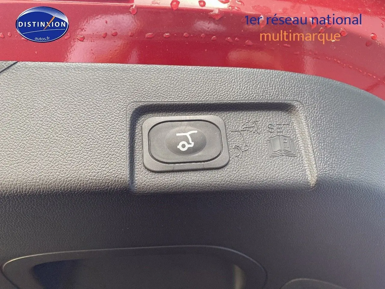 Gros plan sur le bouton d'ouverture électrique du coffre, entouré de plastique noir, sur une Ford Puma rouge.