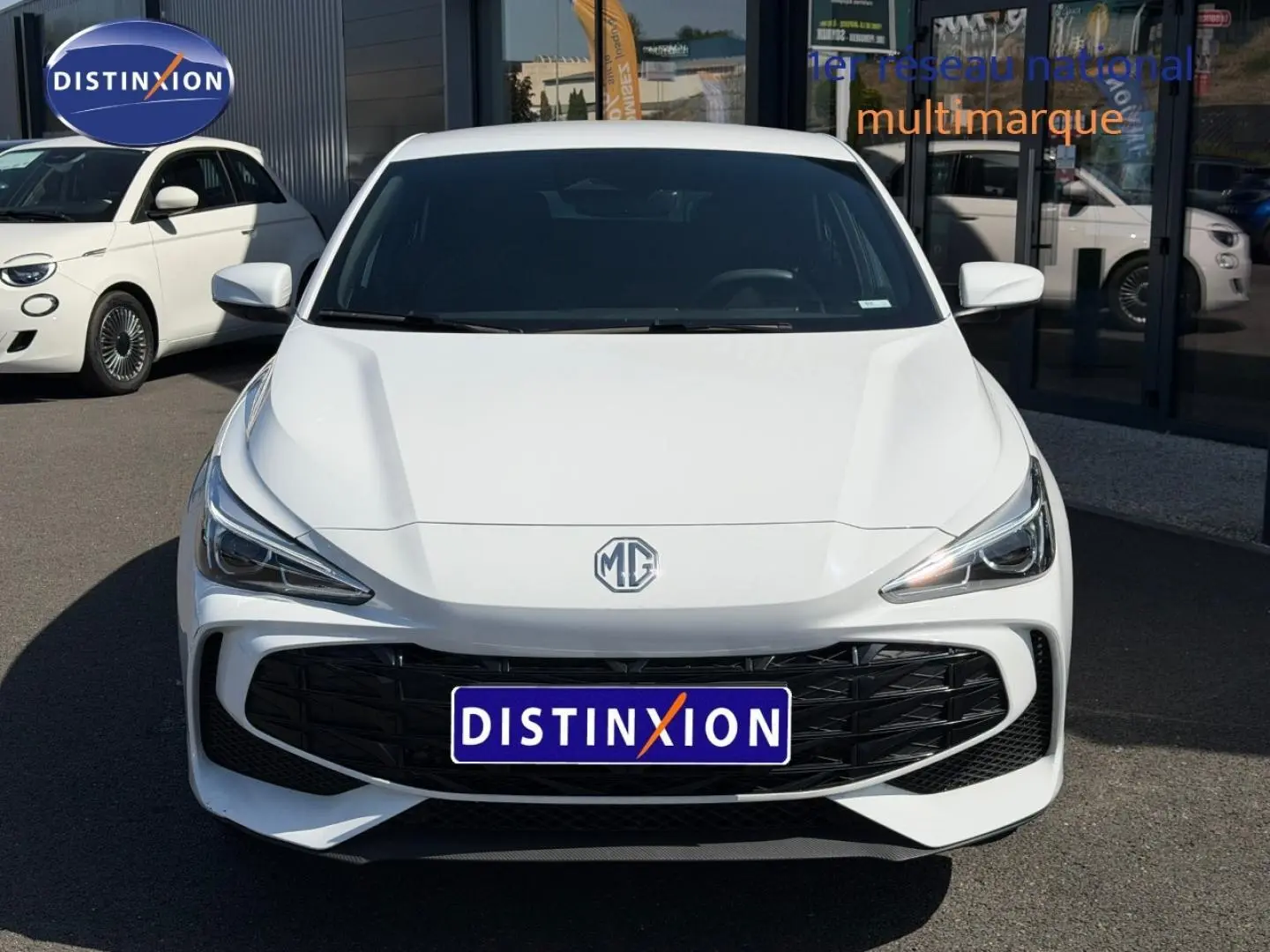 Vue avant d'une MG3 1.5 Hybrid+ blanche avec calandre noire et phares LED, stationnée devant un bâtiment commercial.