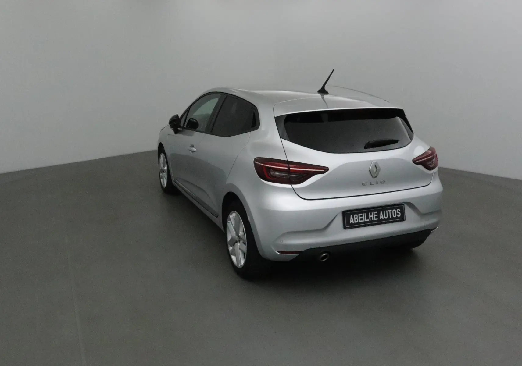 Renault Clio V gris platine vue 3/4 arrière côté gauche dans un studio avec vitres teintées et plaque Abeilhe Autos.