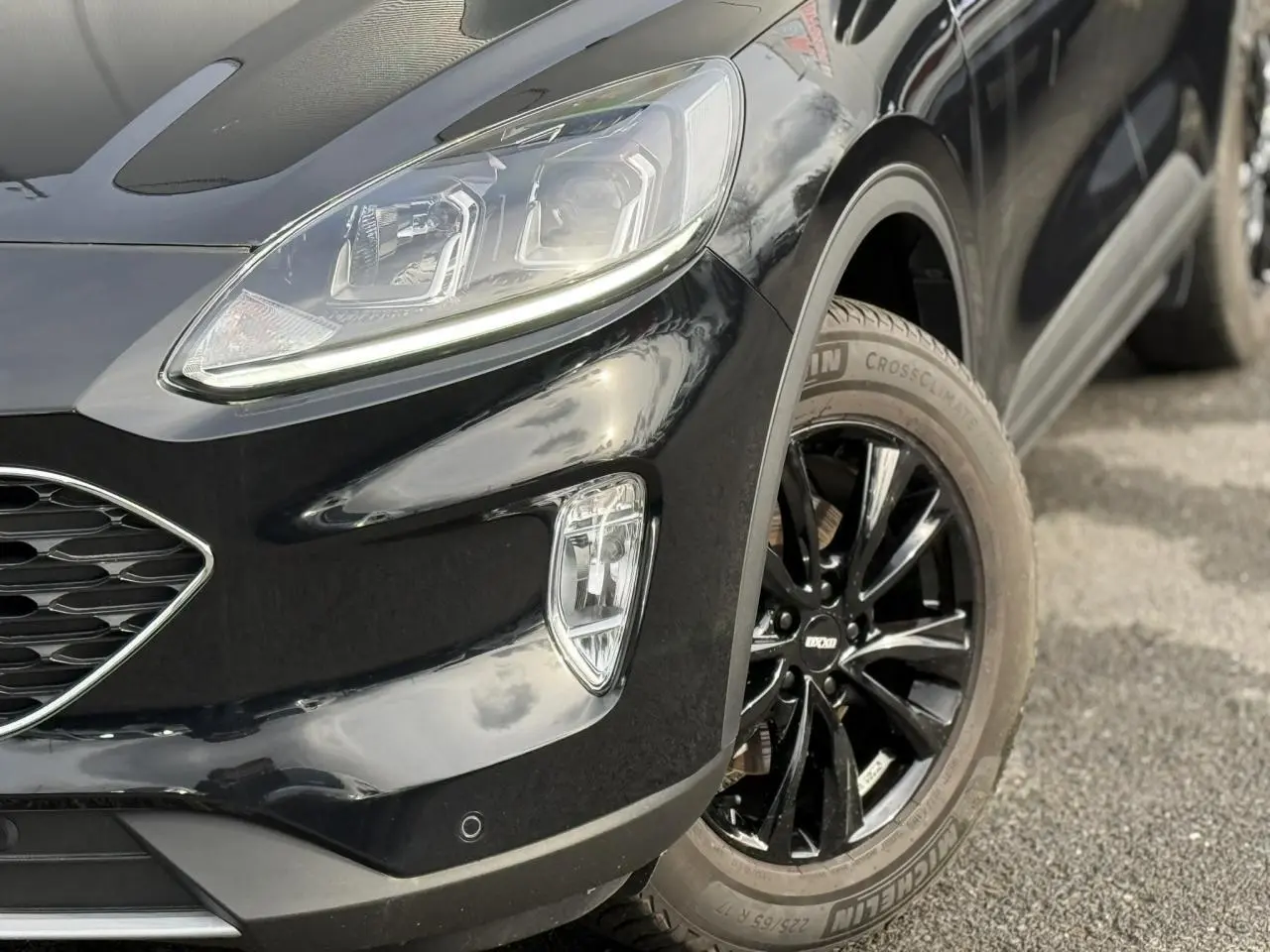 Gros plan sur l'avant droit du Ford Kuga noir Obsidien 2024, mettant en valeur le projecteur et la jante alliage noire.