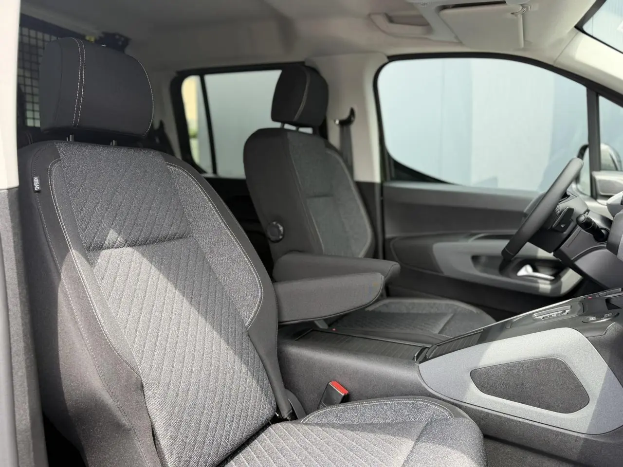 Vue intérieure côté conducteur du Peugeot Rifter 2026, sièges tissu gris avec accoudoir central et volant multifonction.