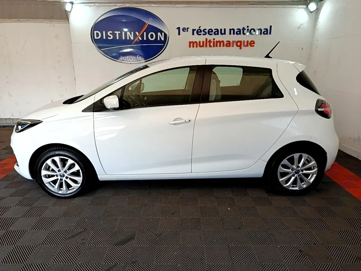 Profil côté gauche de la Renault ZOE Zen R110 blanche, compacte et électrique, sur fond intérieur avec logo Distinxion.