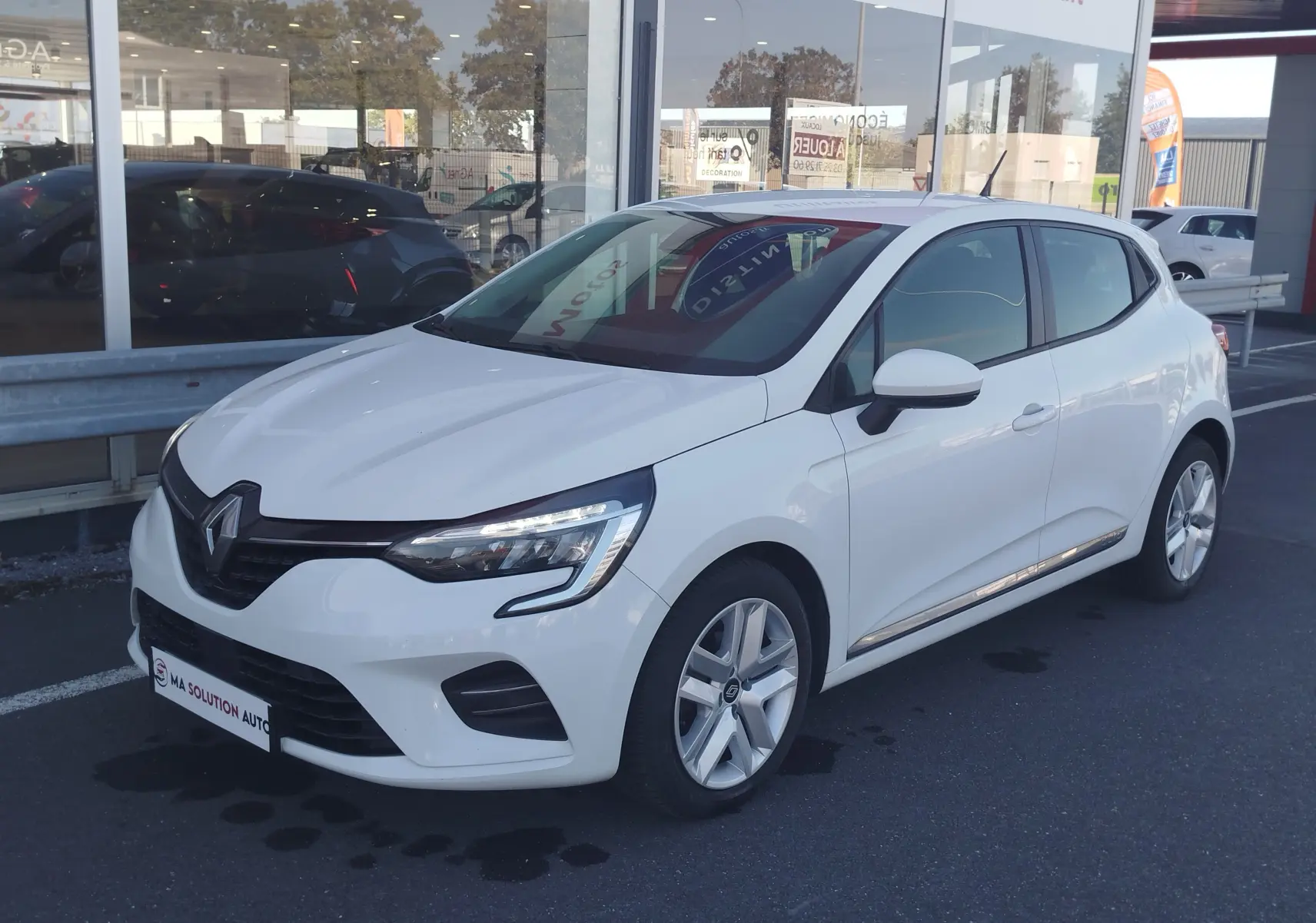 Renault Clio Business blanche vue 3/4 avant droit, avec feux LED et jantes alliage visibles, stationnée devant un showroom.