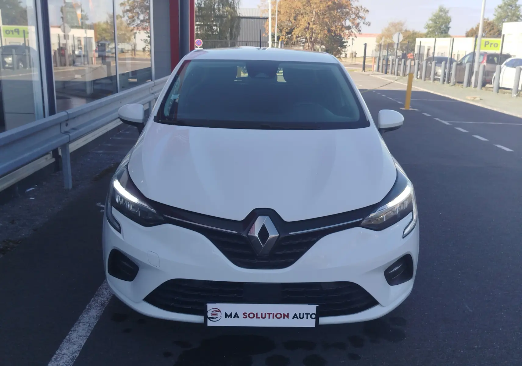 Vue avant d'une Renault Clio Business blanche de 2021 avec feux LED allumés, stationnée devant un bâtiment.