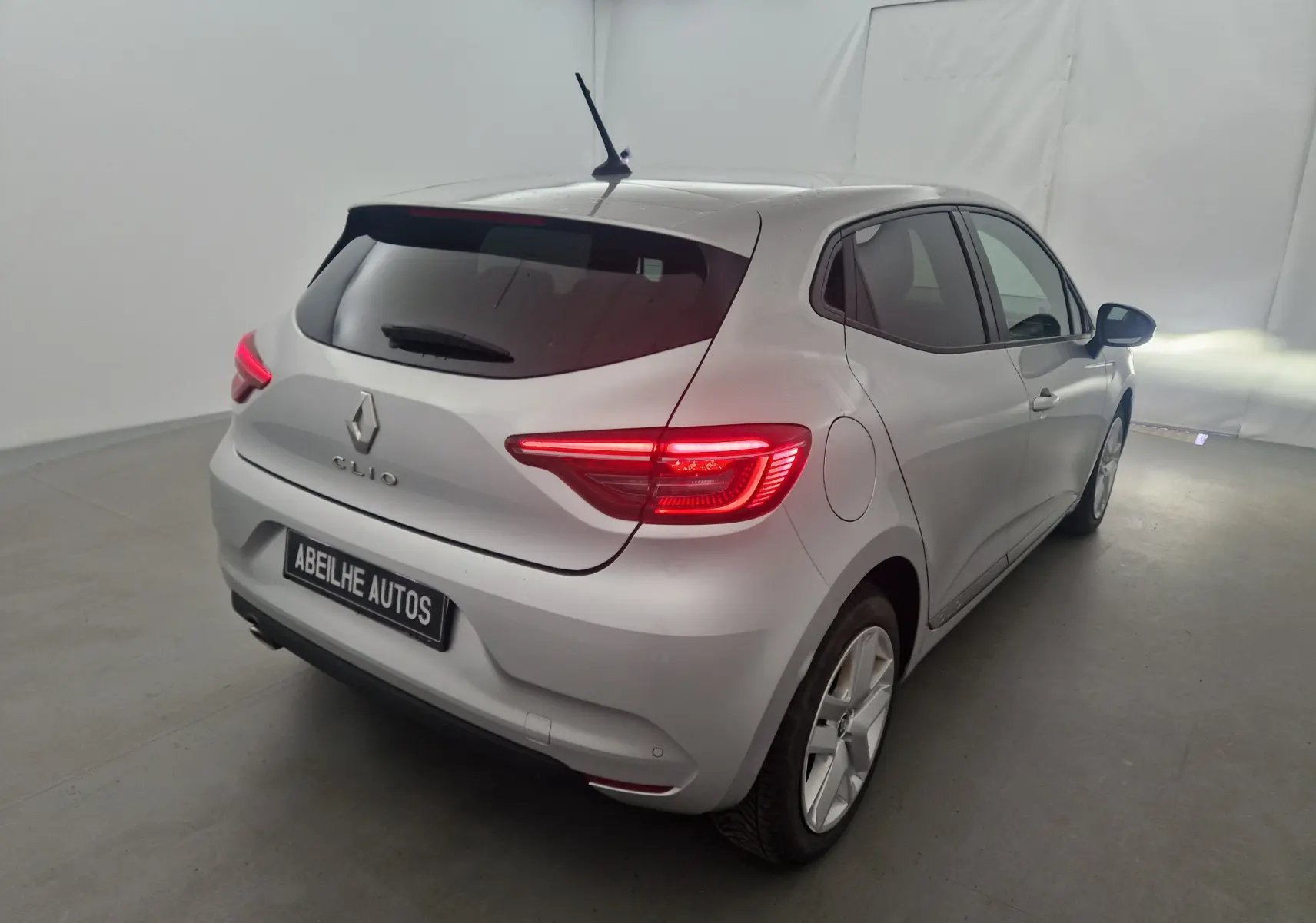 Vue 3/4 arrière droite d'une Renault Clio V gris platine avec feux arrière allumés dans un studio blanc.
