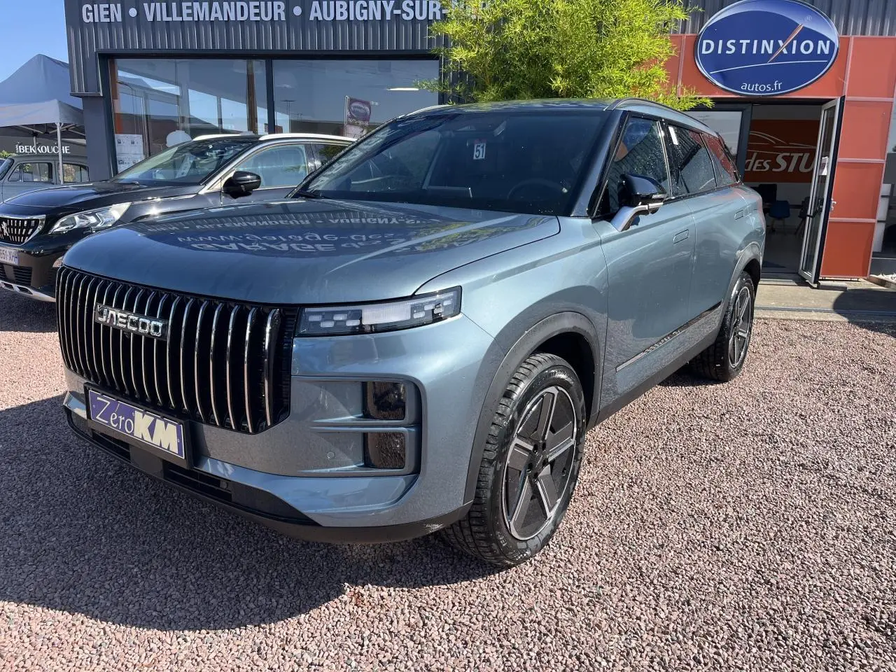 Vue 3/4 avant d'un JAECOO J7 gris olive avec calandre verticale noire et jantes modernes, stationné devant un showroom.
