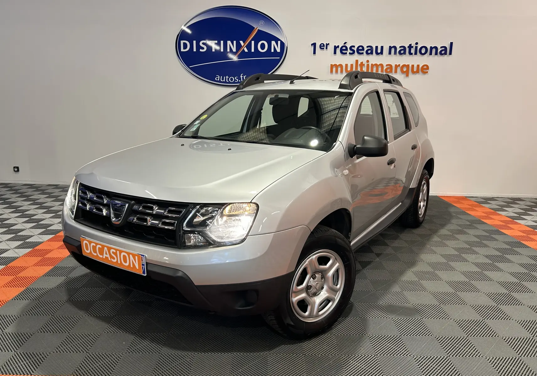 Dacia Duster gris 2017 en 3/4 avant droit avec calandre noire et jantes acier, dans un showroom multimarque.