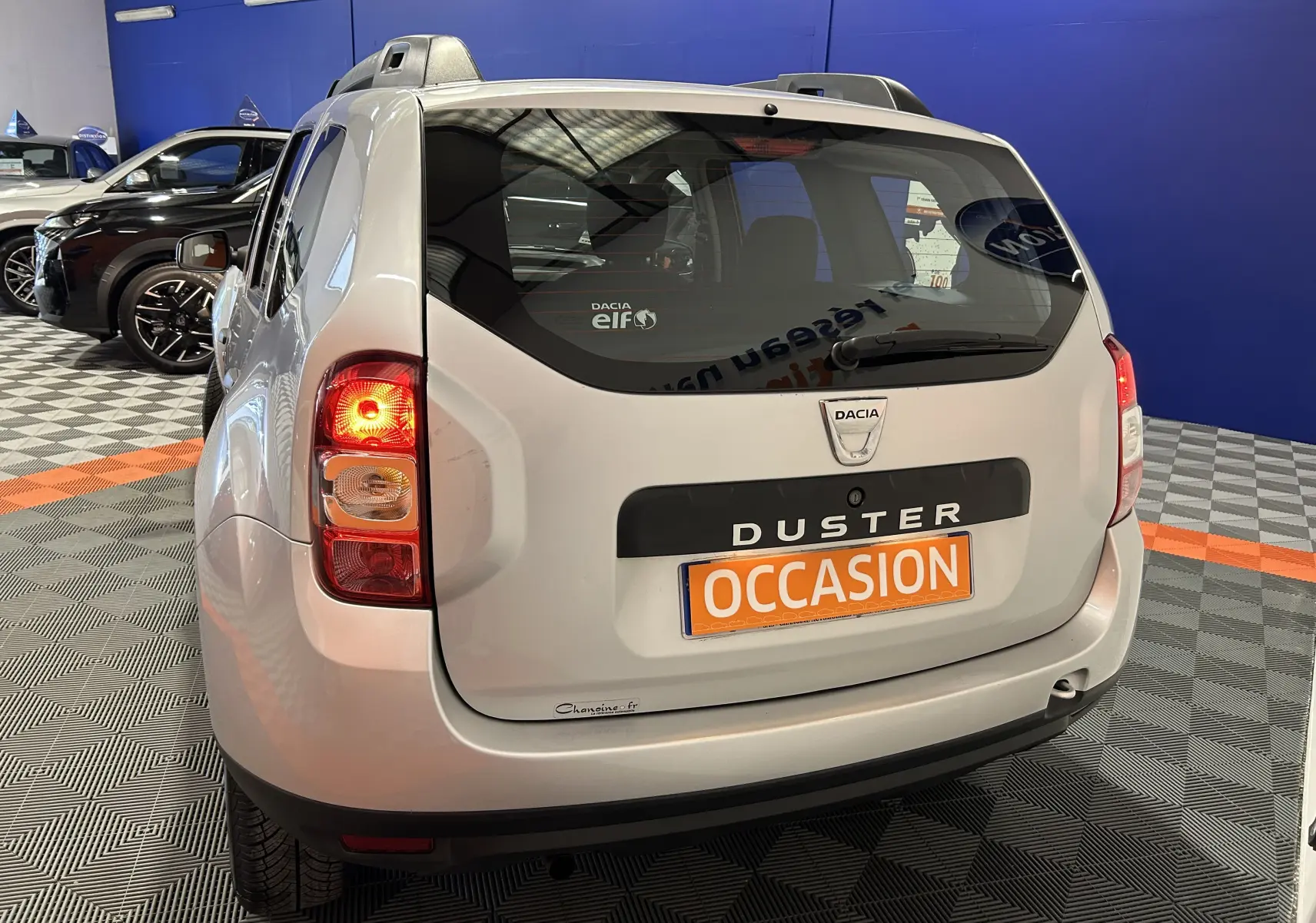 Vue arrière droite d'un Dacia Duster gris 2017 avec feu arrière allumé et plaque "OCCASION" en concession.