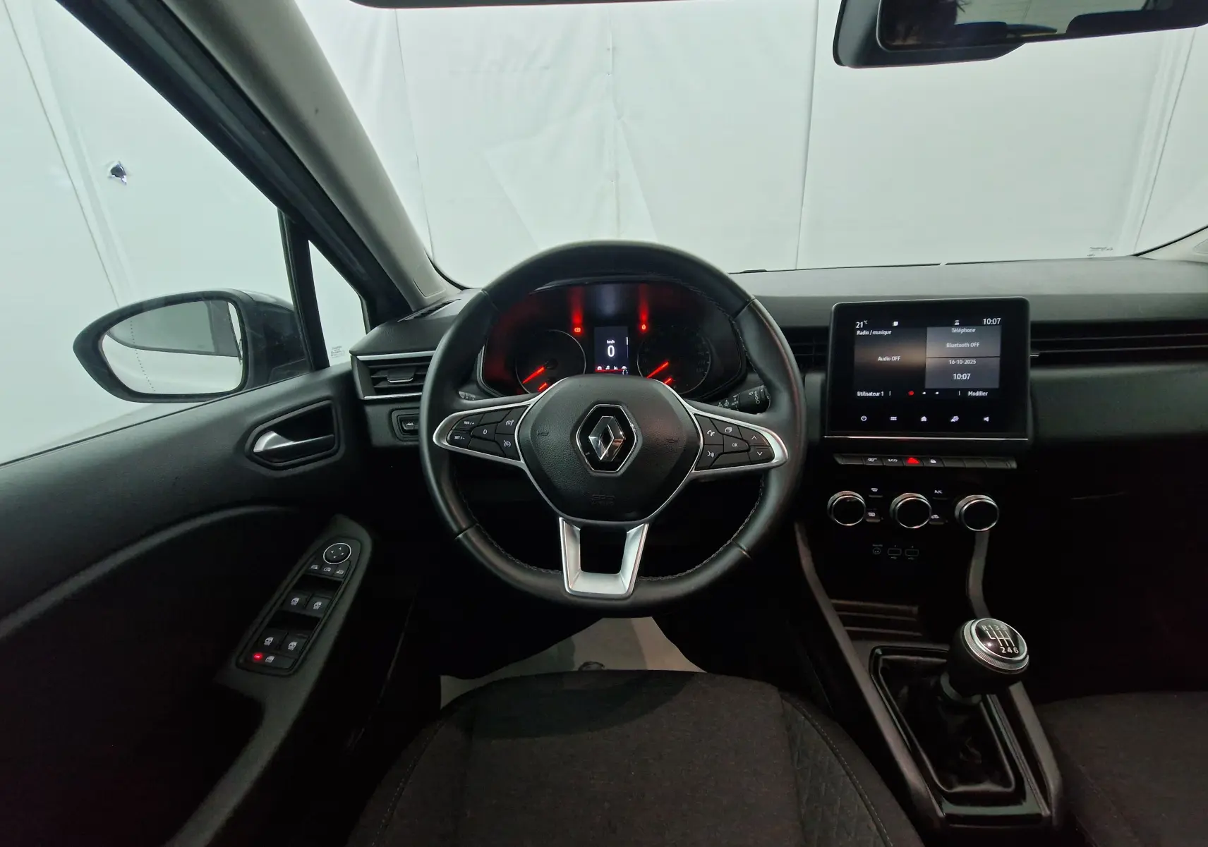 Vue intérieure centrée sur le volant Renault Clio V gris platine, tableau de bord digital et levier de vitesse manuel visible.