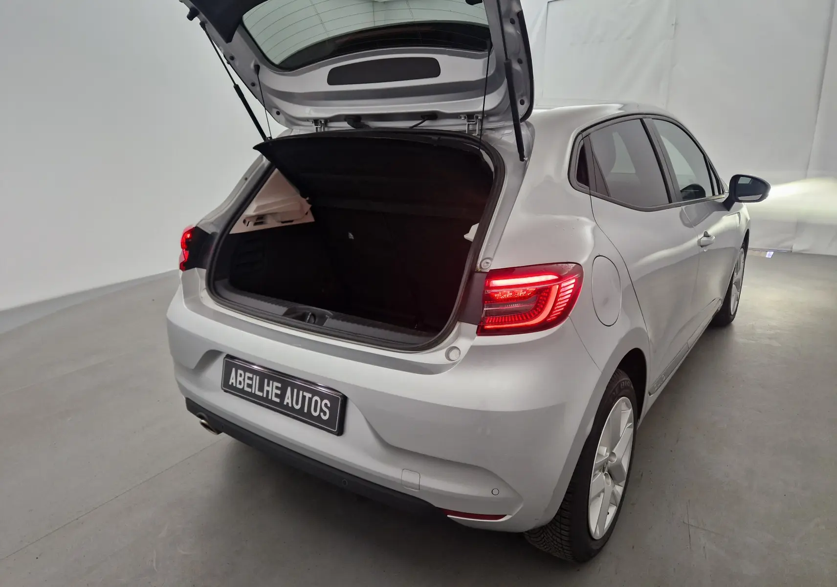 Vue arrière droite d'une Renault Clio V gris platine 2021 avec coffre ouvert et feux arrière allumés.