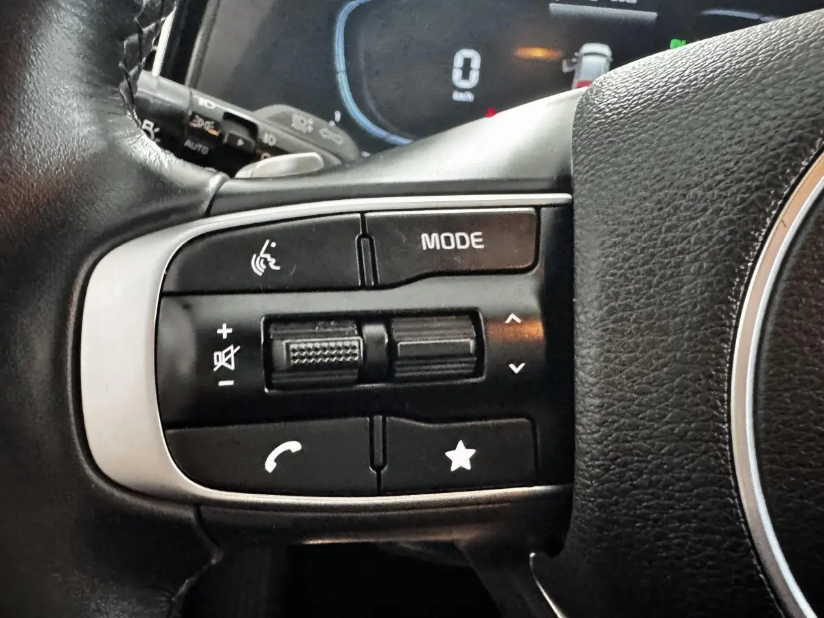 Gros plan sur les commandes gauche du volant cuir noir du Kia Sportage blanc 2024, avec boutons multifonctions.