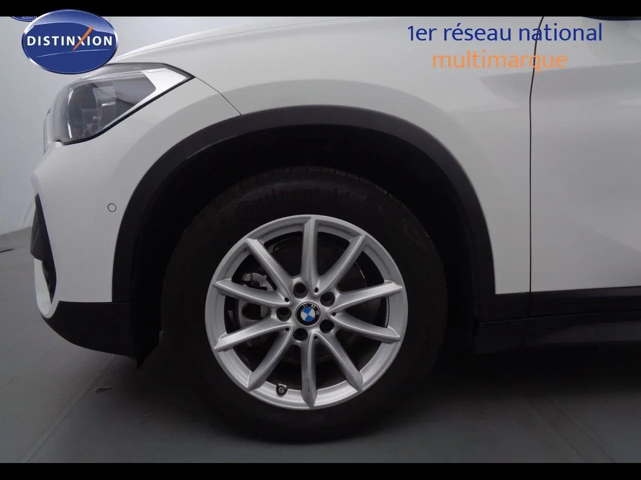 Gros plan sur la roue avant gauche d’un BMW X1 blanc Alpine Metal, mettant en valeur la jante en alliage et le phare avant.