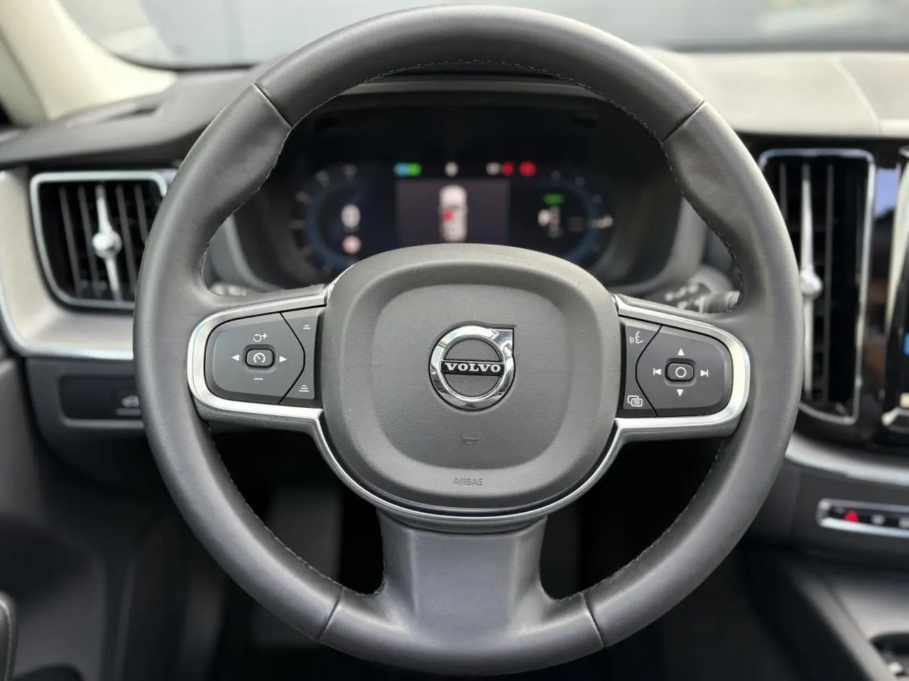 Vue rapprochée du volant simili-cuir noir du Volvo XC60 2024 avec commandes intégrées et tableau de bord digital flou.